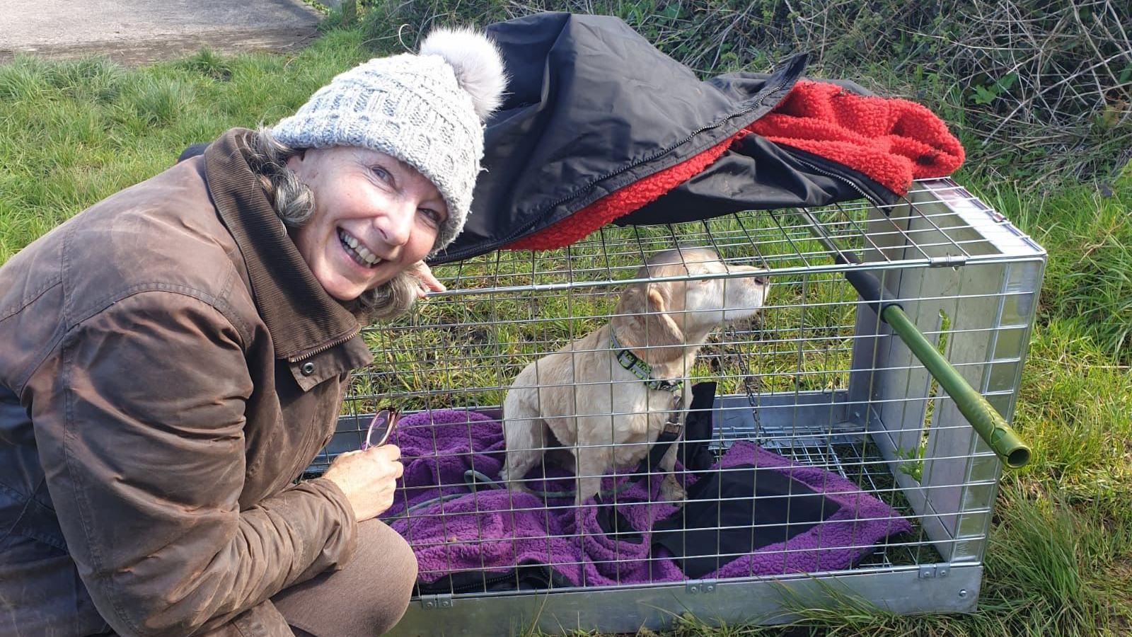 A woman smiles beside a humane cage trap with a puzzled looking labrador dog inside it