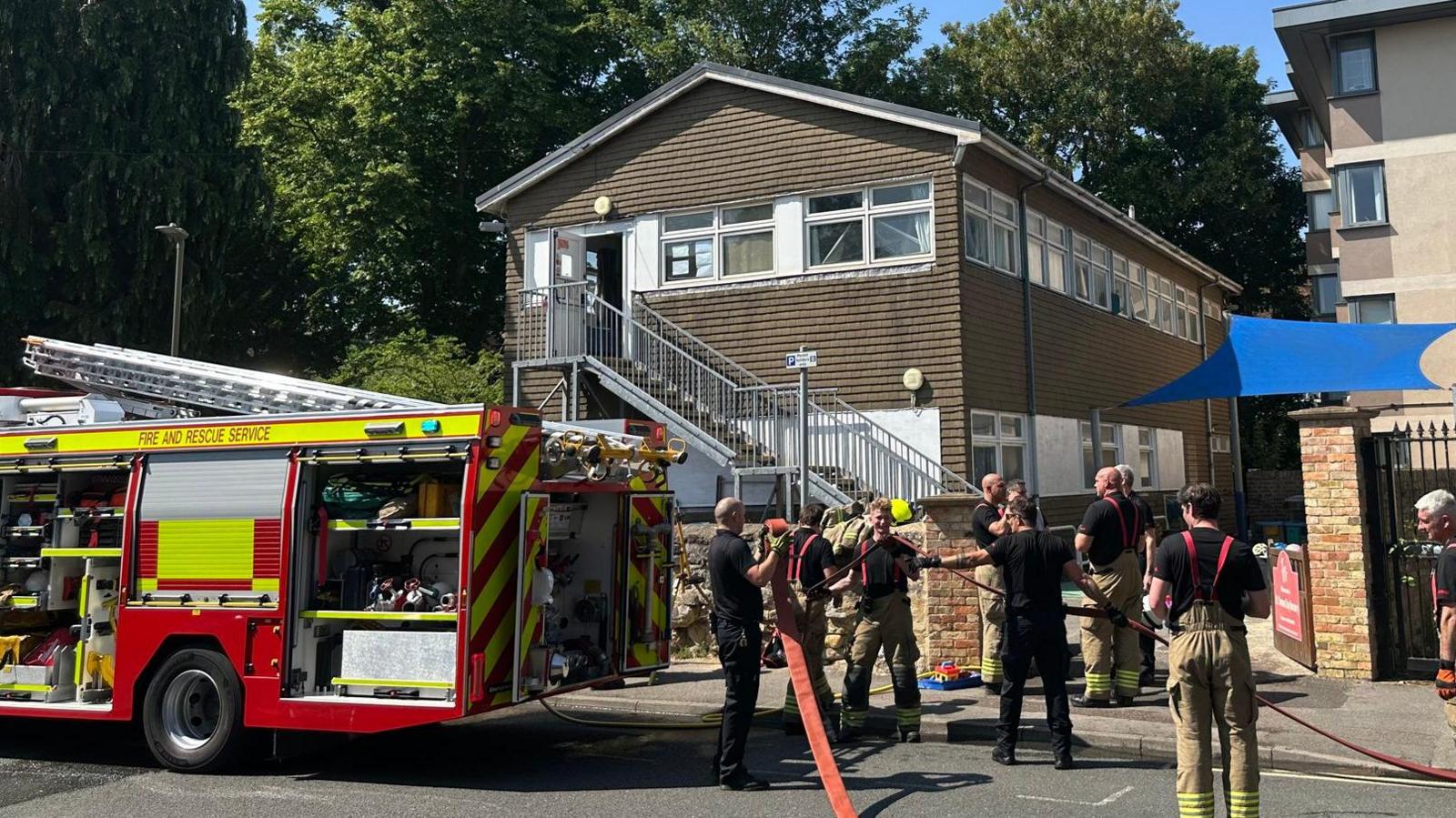 A fire engine and a large group of firefighters stood outside the large brown nursery building.