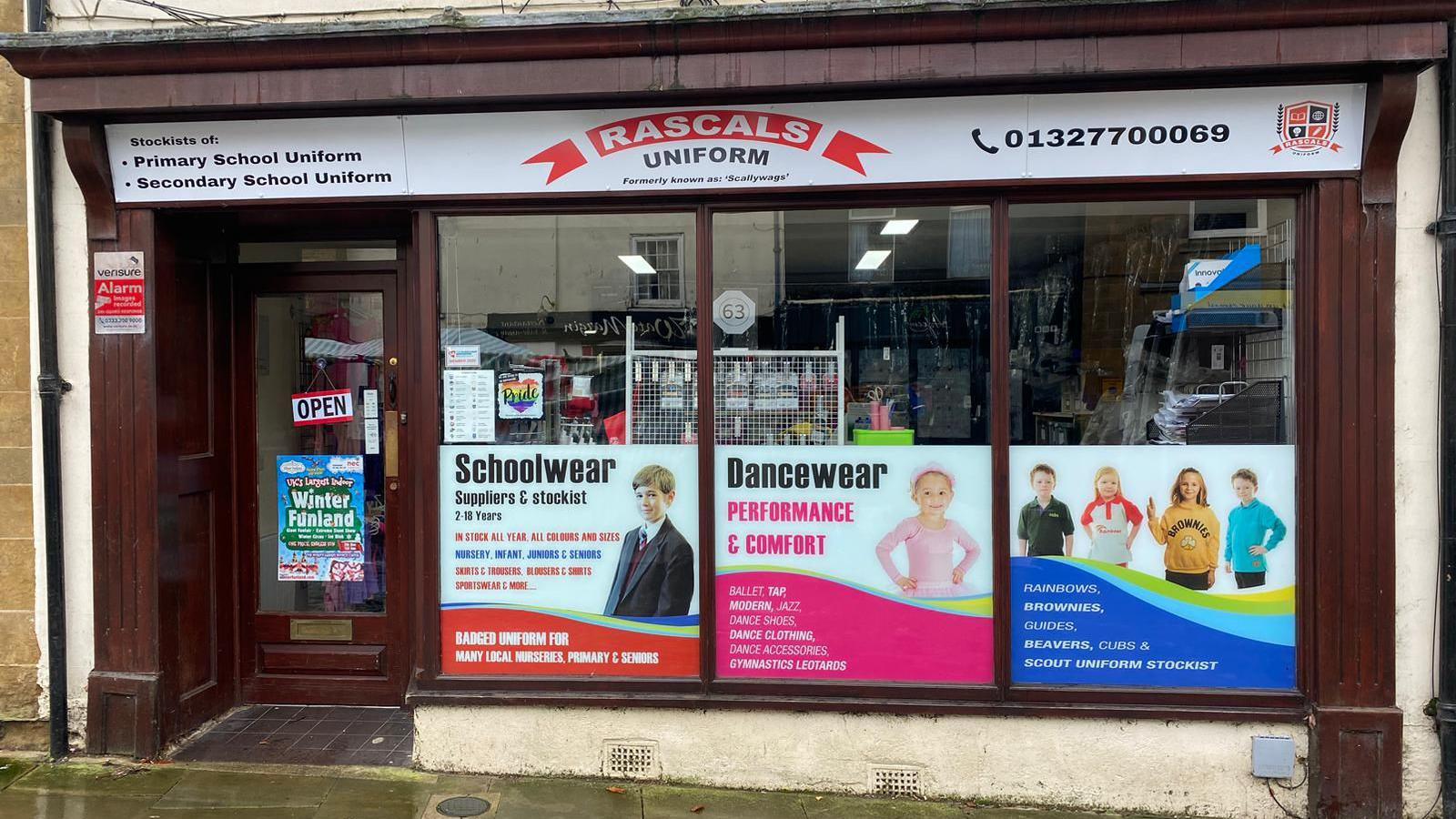 A wooden edged shop frontage with three posters advertising school and dance ware in the windows. The sign for the shop lists a telephone number 