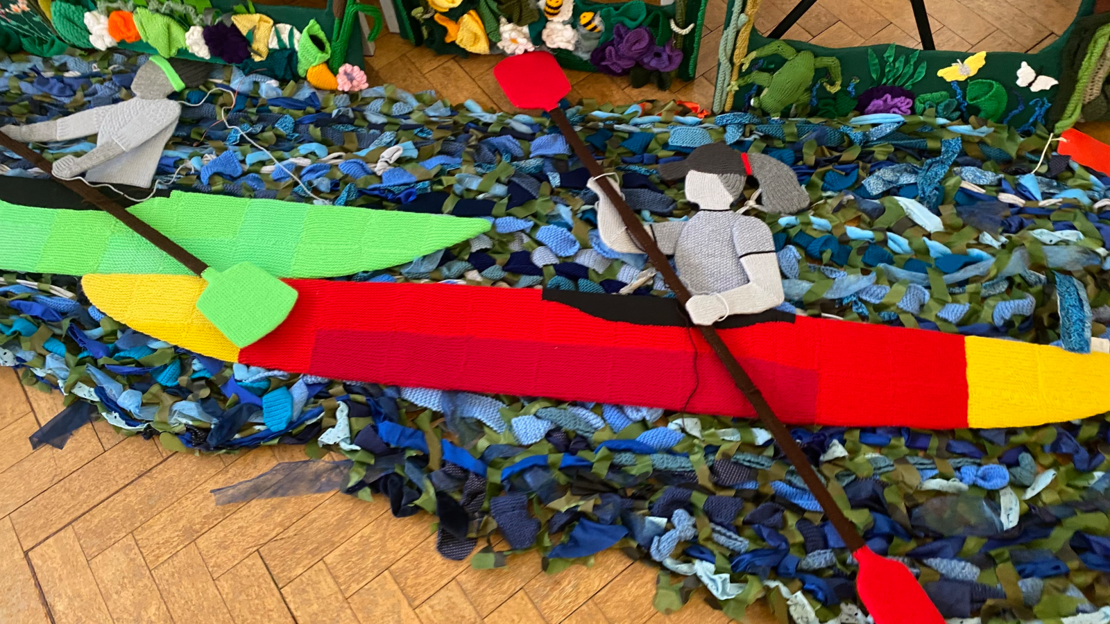 Two kayker's made out of fabric, holding oars, on a river bed, also made out of fabric. The items are placed on a wooden floor. 
