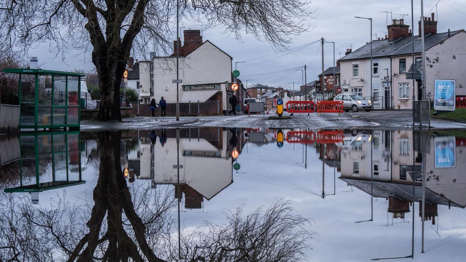 A large area of flooded road with a bus stop to the left. A tree is reflected in the water as well as some buildings and an orange road block. 