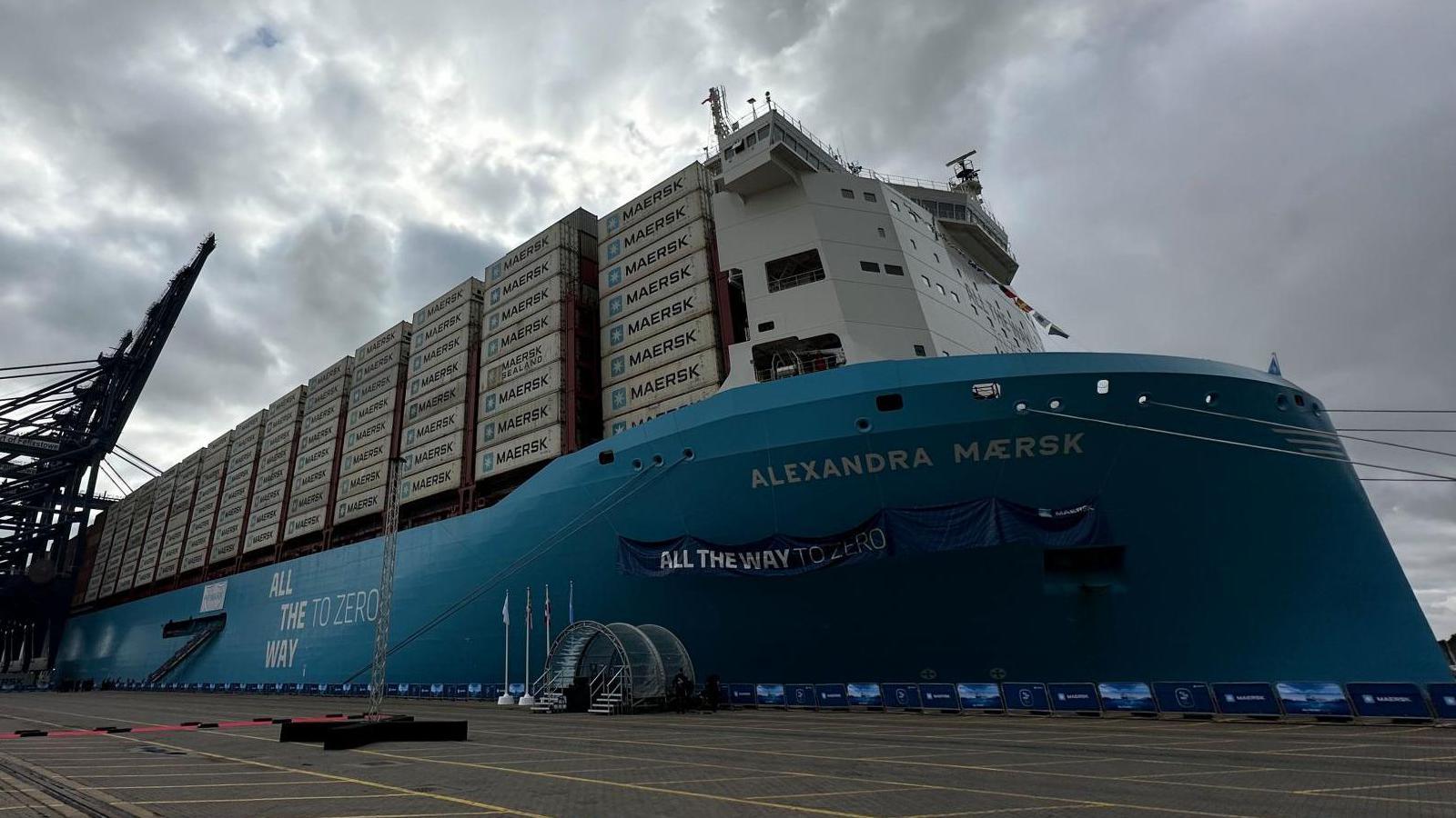 Large container ship moored next to some cranes. The ship has a large number of containers onboard. It has a blue hull. 