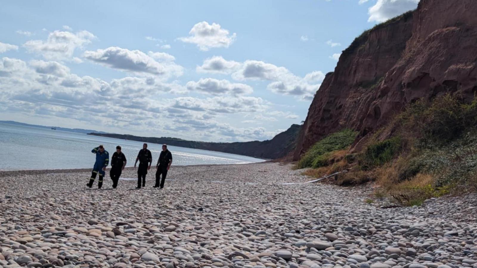 Coastguard teams walking along Budleigh Beach. There is a cliff on the right and it is a pebble beach. The sea is on the left.