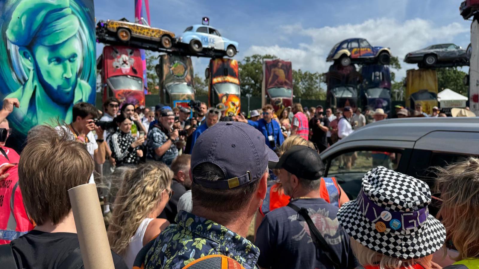 The roof of a car with Michael Eavis' in, surround by festival goers. On the left is a green and black portrait of Eavis and in background is more of Carhenge.