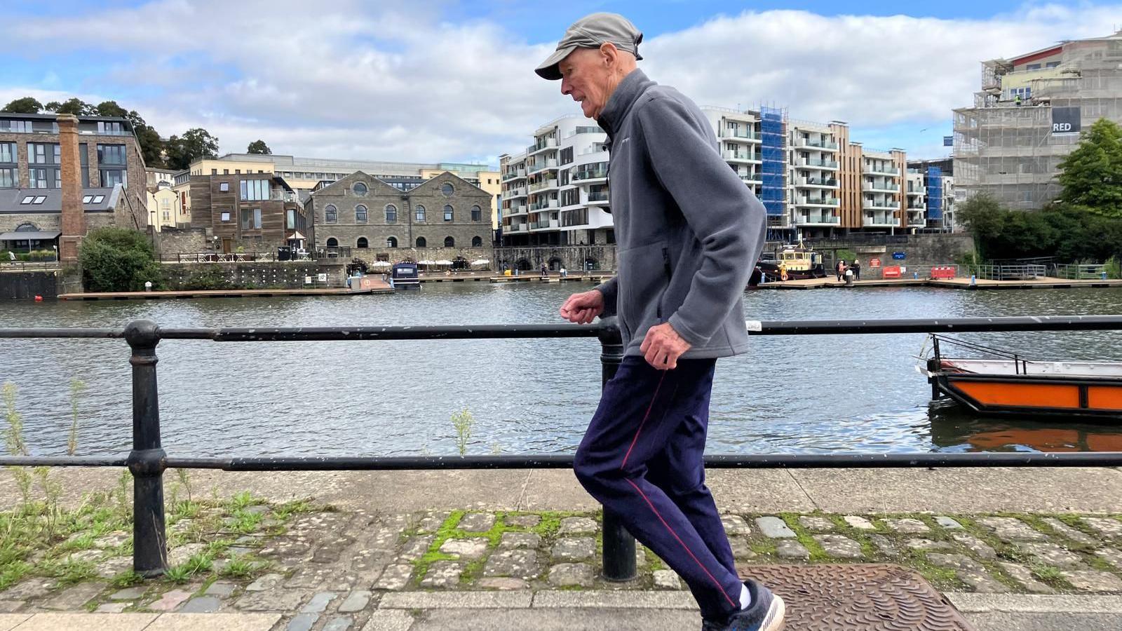 92-year-old Ken Head is pictured running along Bristol's harbourside. There is a barrier behind him, going along the water's edge, buildings behind and a boat on the river.