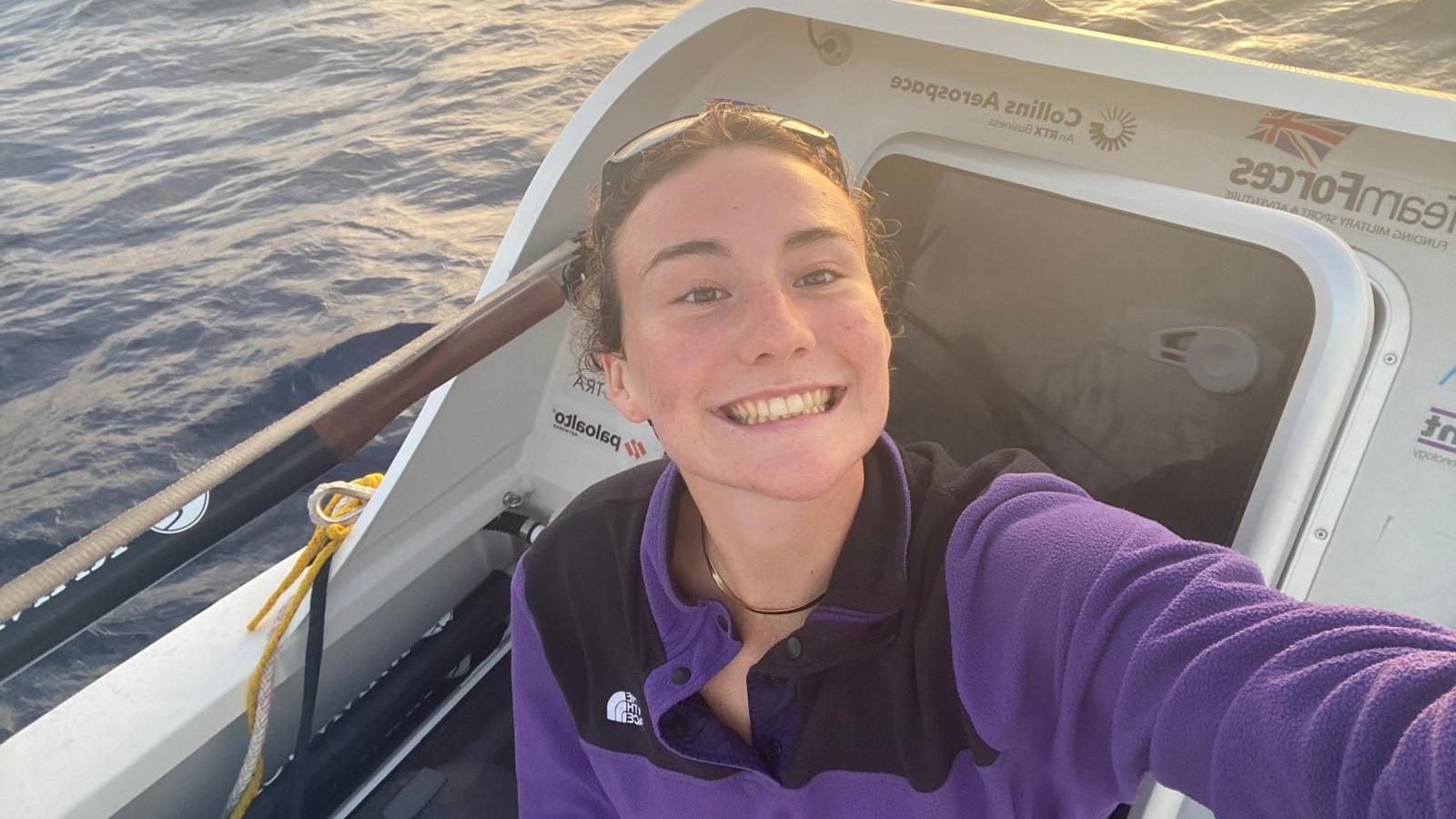 Zara Lachlan wears a purple fleece while on her boat in the ocean. She has a gold chain around her neck and is smiling at the camera. Her brown hair is up and there are sunglasses on her head. 