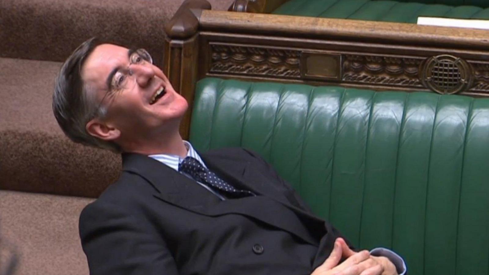 Jacob Rees-Mogg wearing glasses, smiling and reclining on one of the green leather benches in the House of Commons