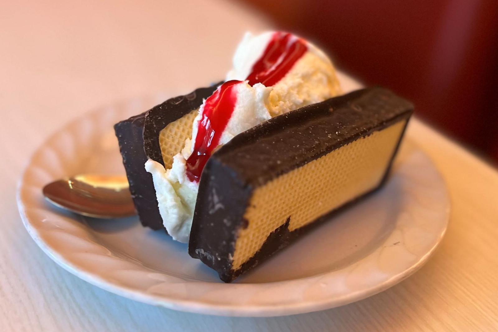 Two scoops of ice cream covered in sauce are sandwiched between chocolate covered, mallow-filled wafers. The treat sits on a white plate on a table.