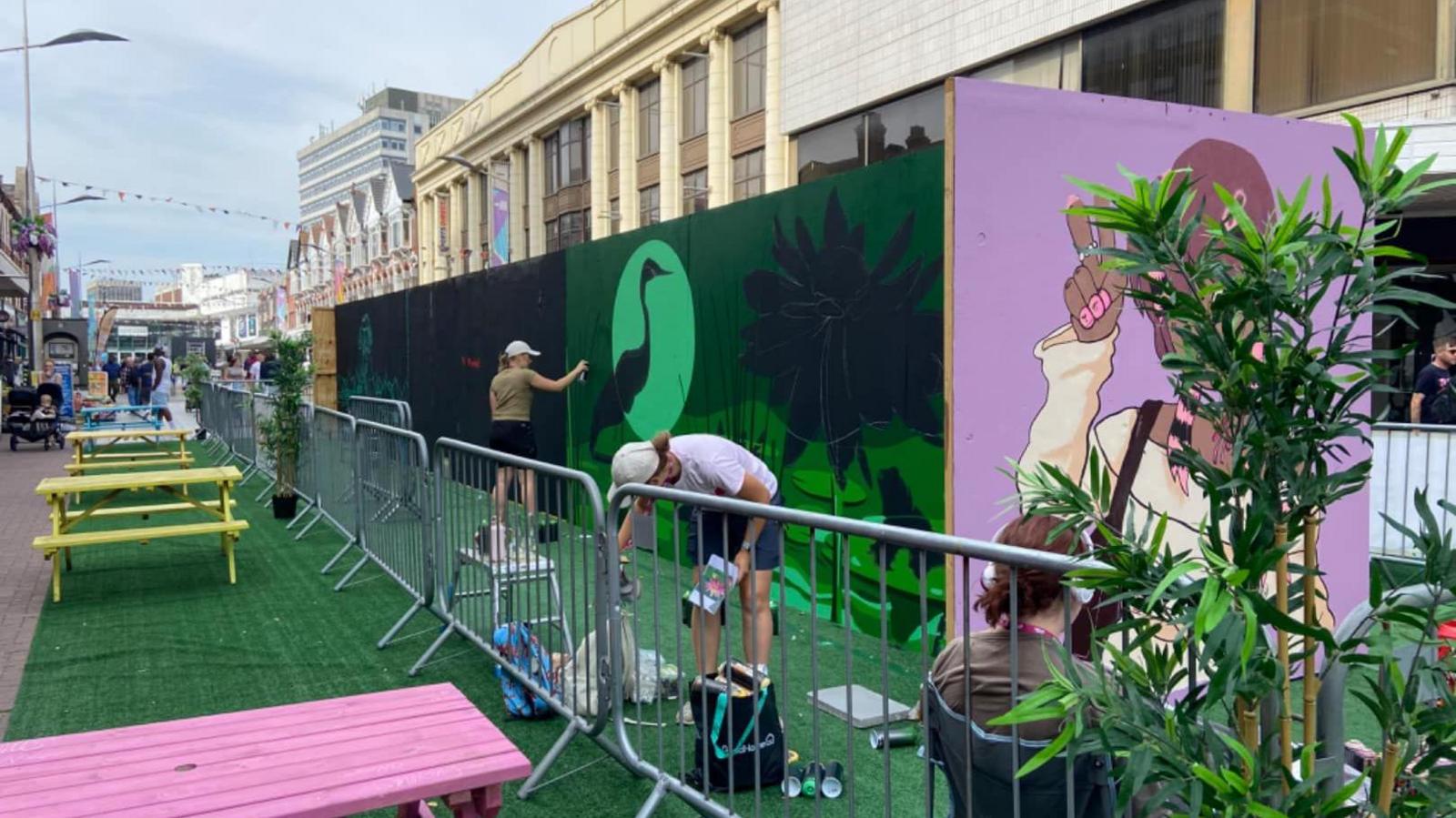 Two artists creating artwork on temporary wooden walls in a Southend street. One of them is spray painting black paint.