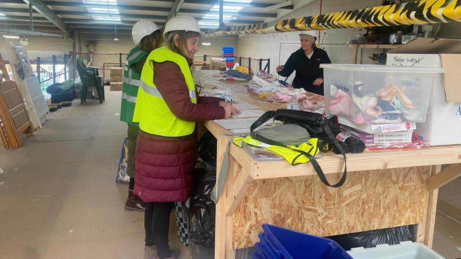 Three women in an industrial unit sorting out clothing. There are lots of baby items on a wooden desk, and the women are all wearing white hard hats. The woman at the front of the image has on a yellow high-vis jacket, and a red coat, another lady to the left has a green high-vis on, and a woman to the right is wearing a black jacket. 