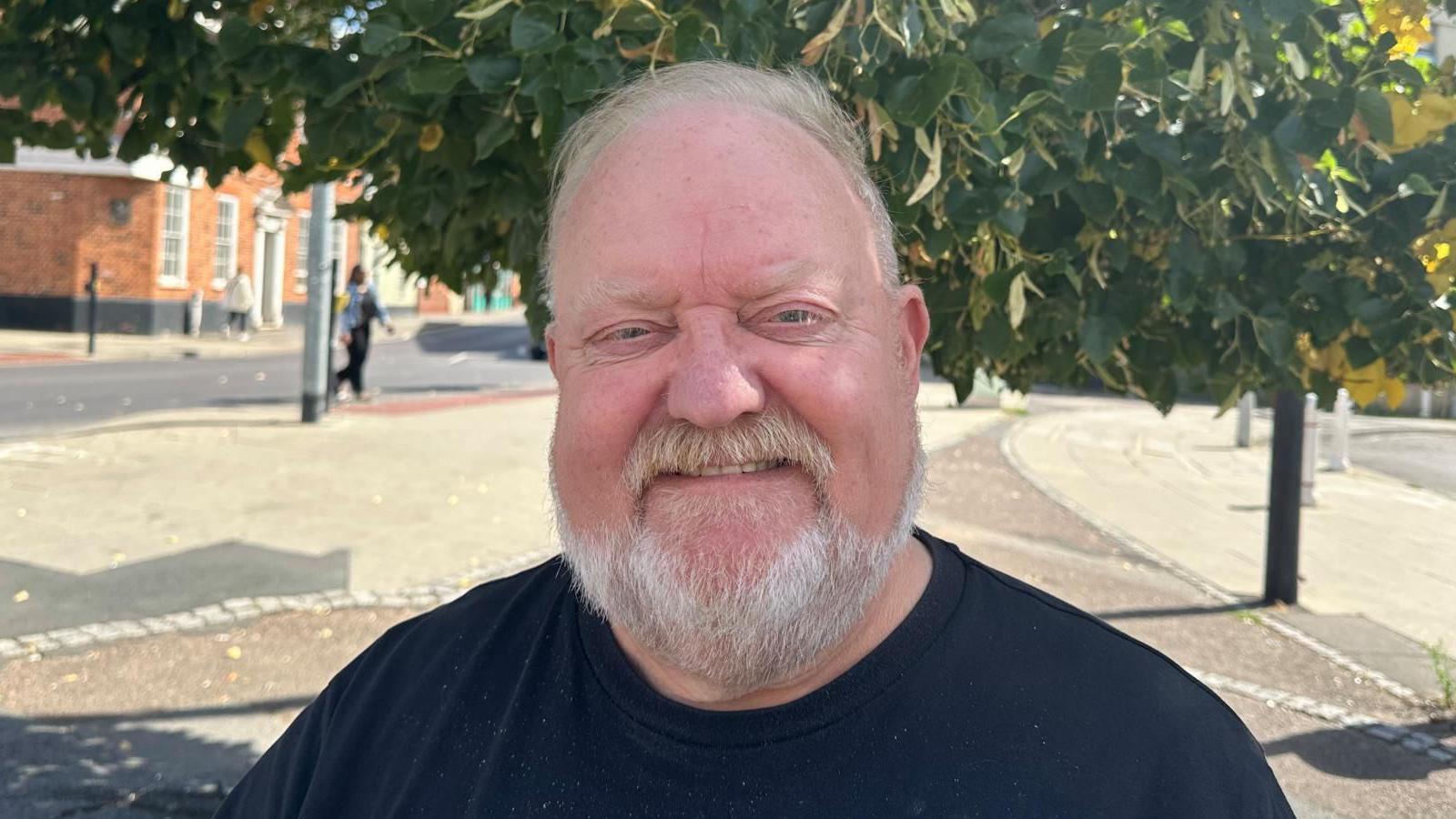 Colin Kreidewolf standing in front of a tree, he has a grey beard and hair, he is wearing a black T-shirt.