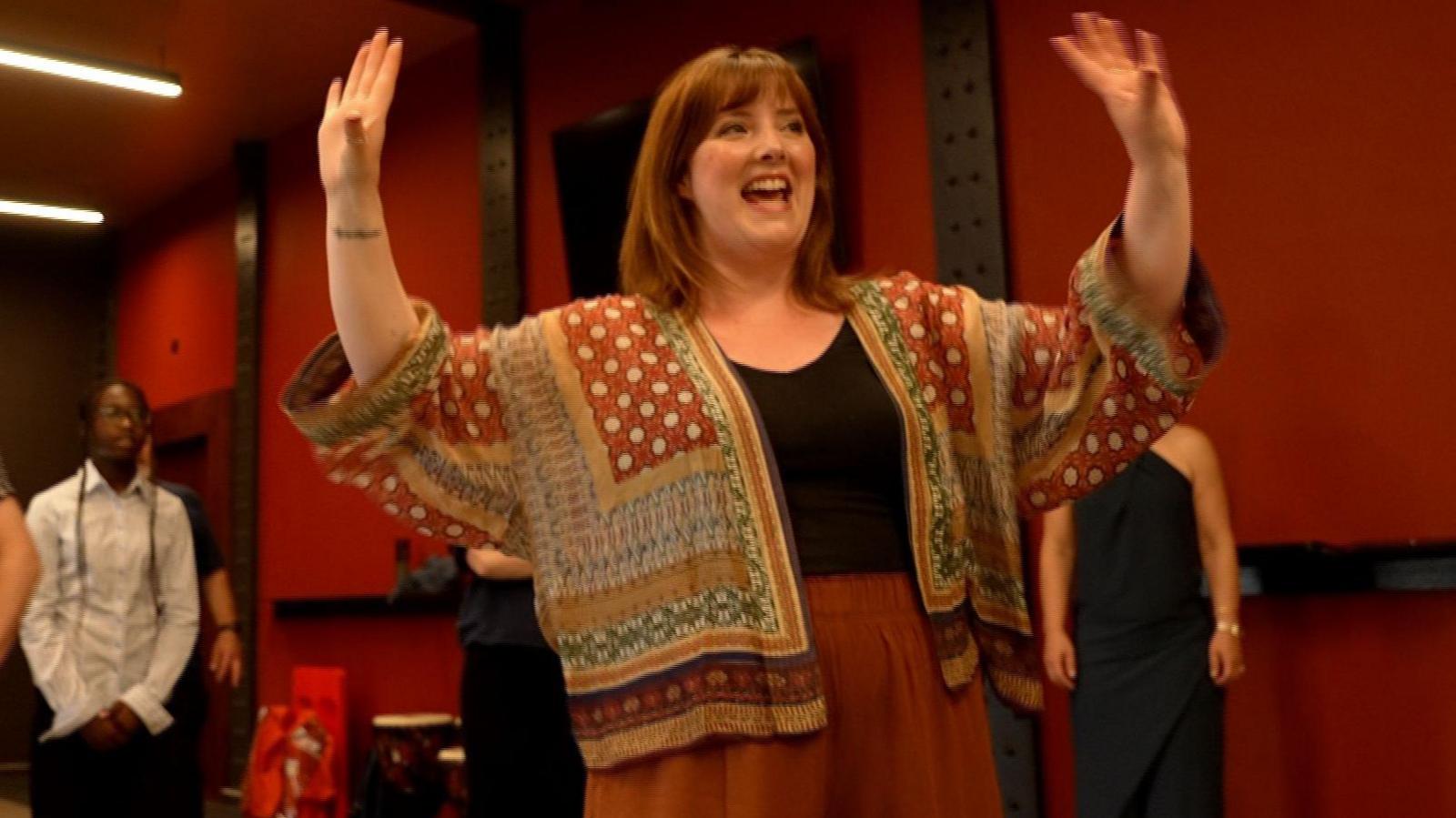 Marie Claire Breen, vocal coach at Opera North in bright patterned red and mustard jacket with black top and arms up, going through vocal warm up before the rehearsal