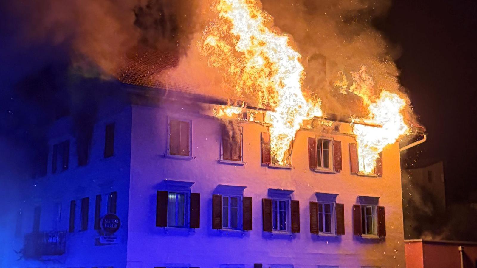 Large flames come out of the windows in the top floor of a building at night.