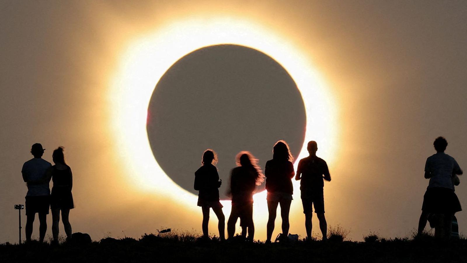Full eclipse of the sun with its corona glow. People are standing in silhouette on high ground.