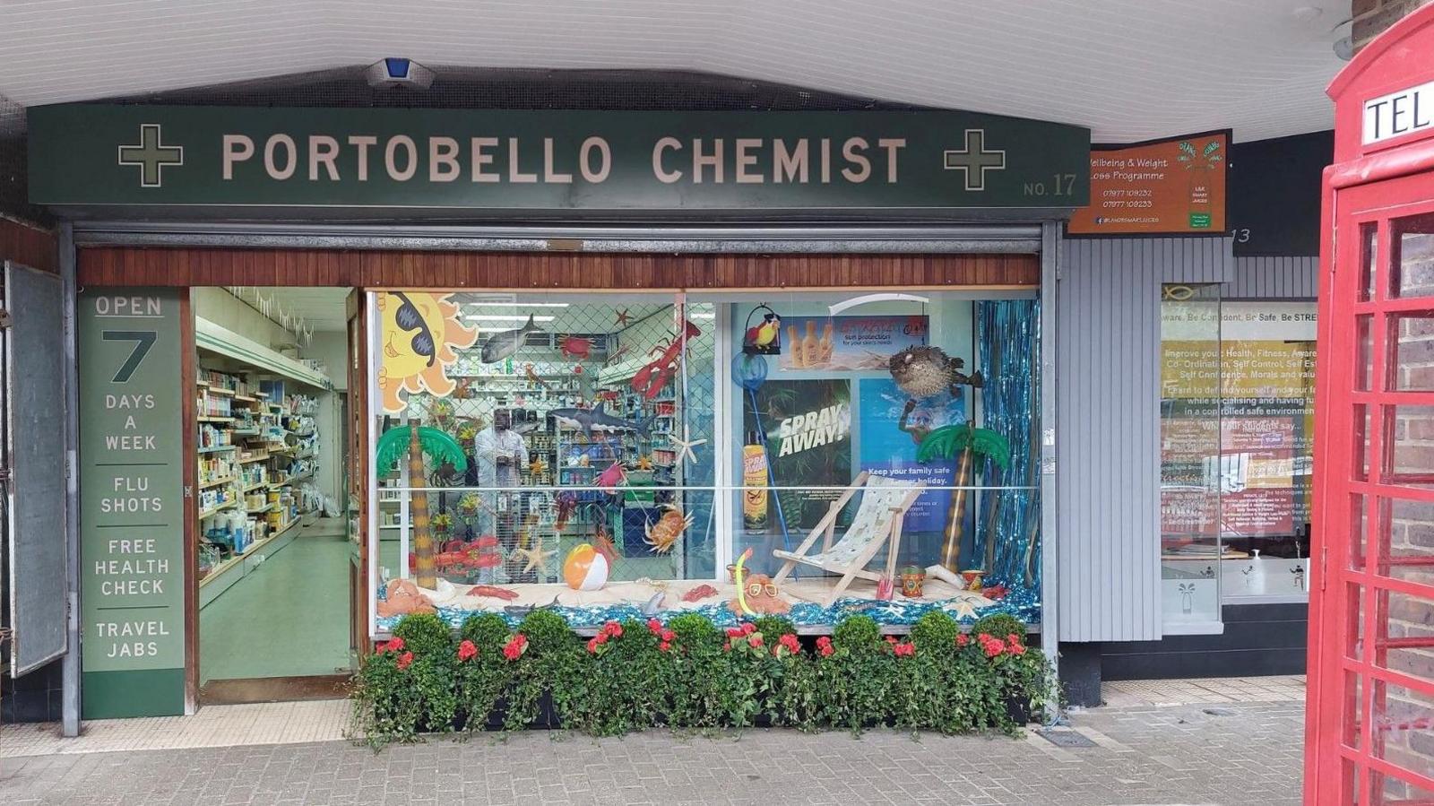 Gadebridge Chemist in Hemel set dressed as "Portobello Chemist" with those words written as the main sign. There is the edge of a red phone box in the picture and the chemist has summery items in the window including a yellow snorkel, white deckchair, beach ball and a poster of a sun wearing sunglasses. In front of the shop are trailing plants with red flowers.