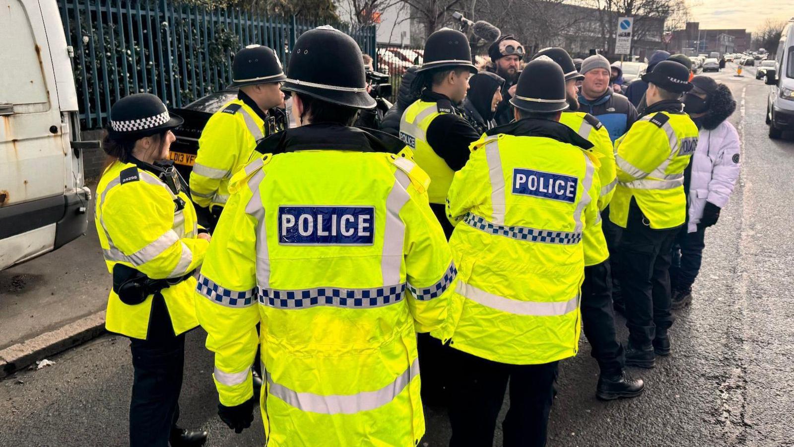 Police officers crowing around protesters on a street. They wear high-vis green jackets with the word police in white letters on the back. Most of the officers wear helmets. There is a group of protesters beyond the officers.