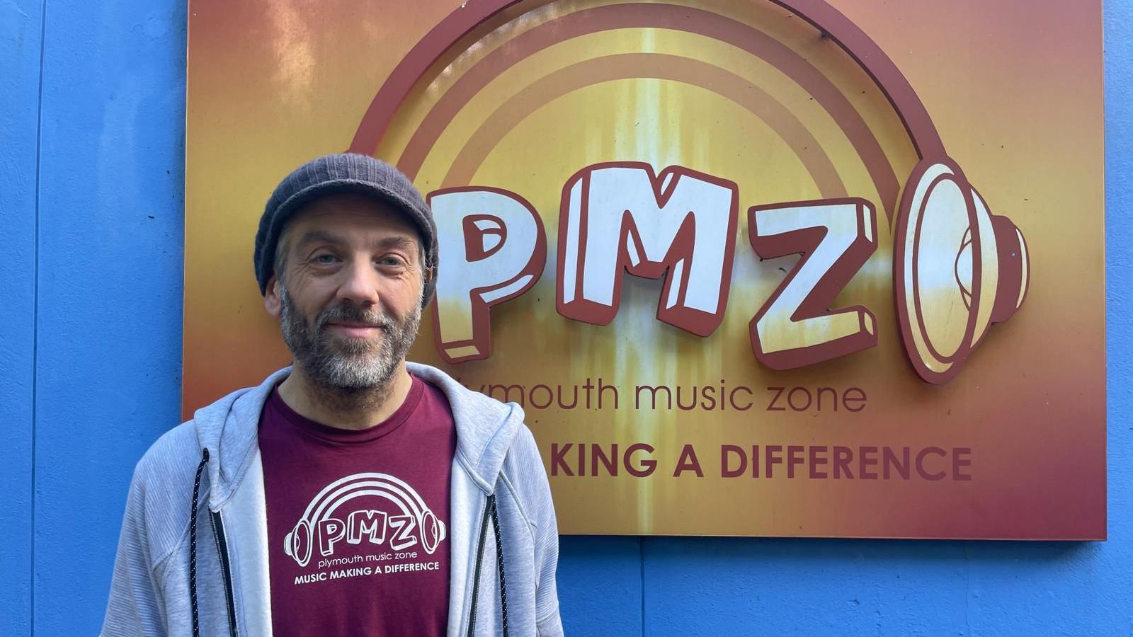 Karl Meyer, who runs Plymouth Music Zone, is pictured infront of an orange sign which has the Plymouth Music Zone logo on it. The logo is a picture of animated headphones with PMZ in the middle. Below it it reads 'Plymouth Music Zone and below that it reads 'Making a difference'.Karl is wearing a burgundy  T-shirt with the same logo on it and a grey hoodie on top.