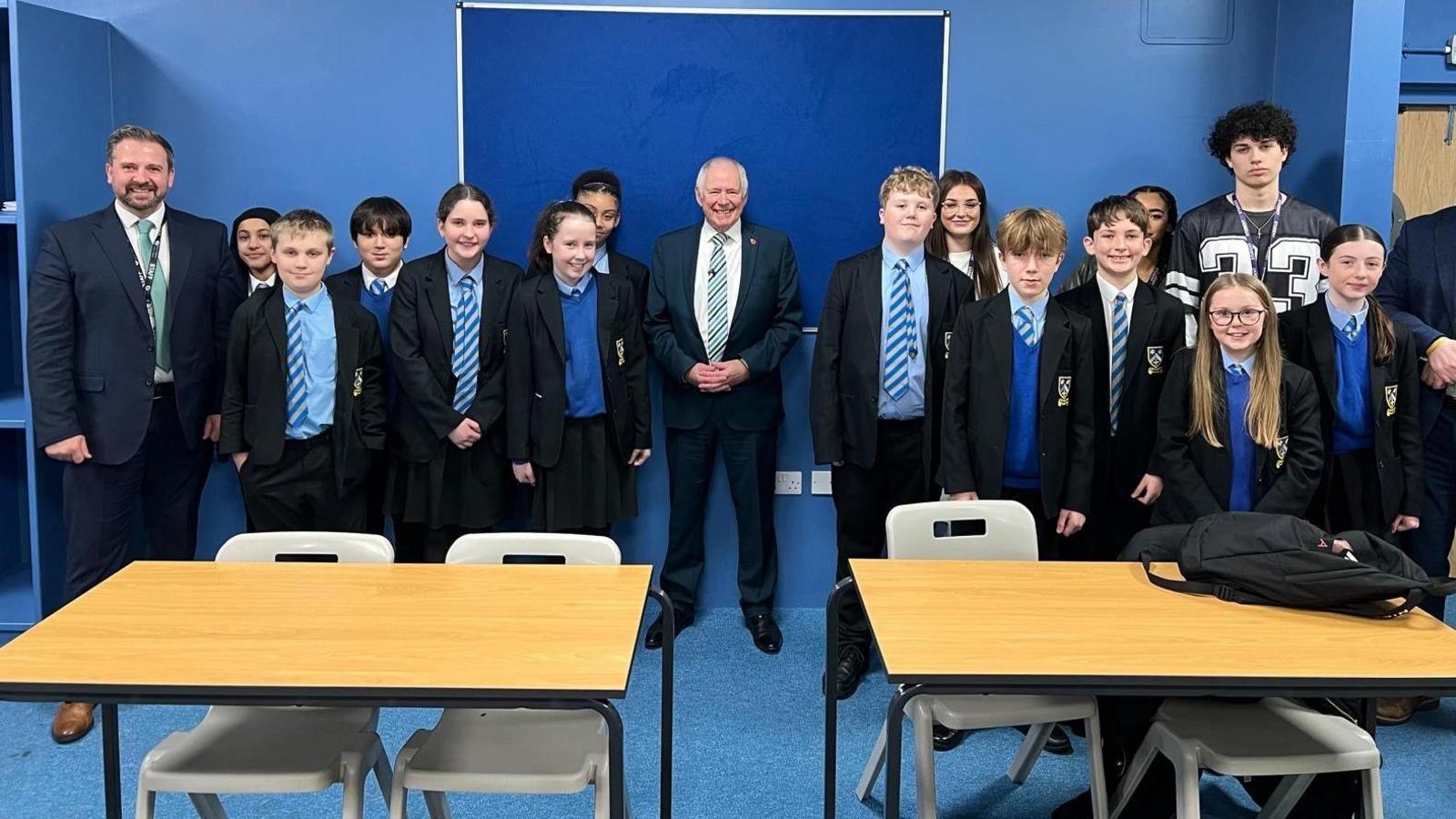 A group of secondary school students stand in a classroom with two adults. The students are wearing school uniforms that consist of black blazers, pale blue shirts, blue jumpers and blue striped ties. There is one man wearing a suit on the left side of the group. There is also a man wearing a suit in the centre of the group. The room has blue walls and pale wooden desks.