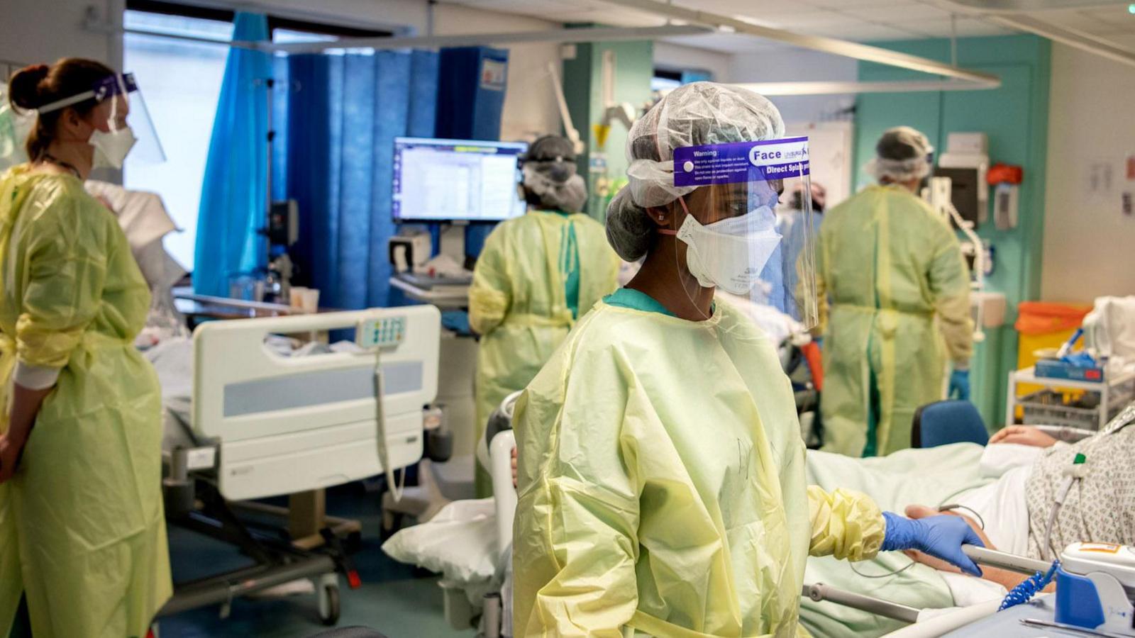 A photo of doctors wearing yellow scrubs over their blue uniform as well as masks and PPE face shields at Addenbrooke’s during Covid. The room is busy and there are several patients lay in hospital beds separated by curtains. 