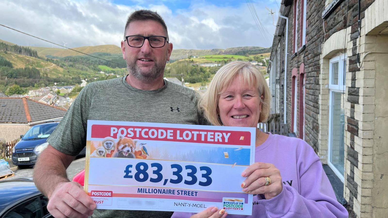 Rob and Michaela Jones on the street holding their winning PostCode Lottery cheque. Rob has short black hair and black glasses on, he is wearing a khaki t-shirt. Michaela has short blonde hair and is wearing a lilac jumper.