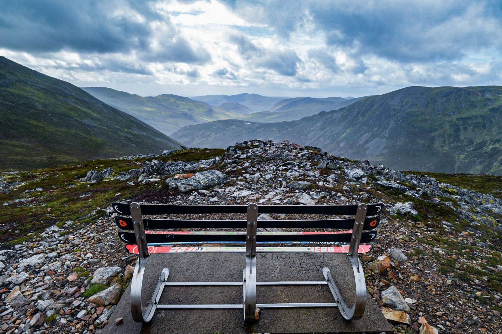 Bench made of ski's at the top of a rocky mountain with other mountains in the distance, below a grey and blue cloudy sky