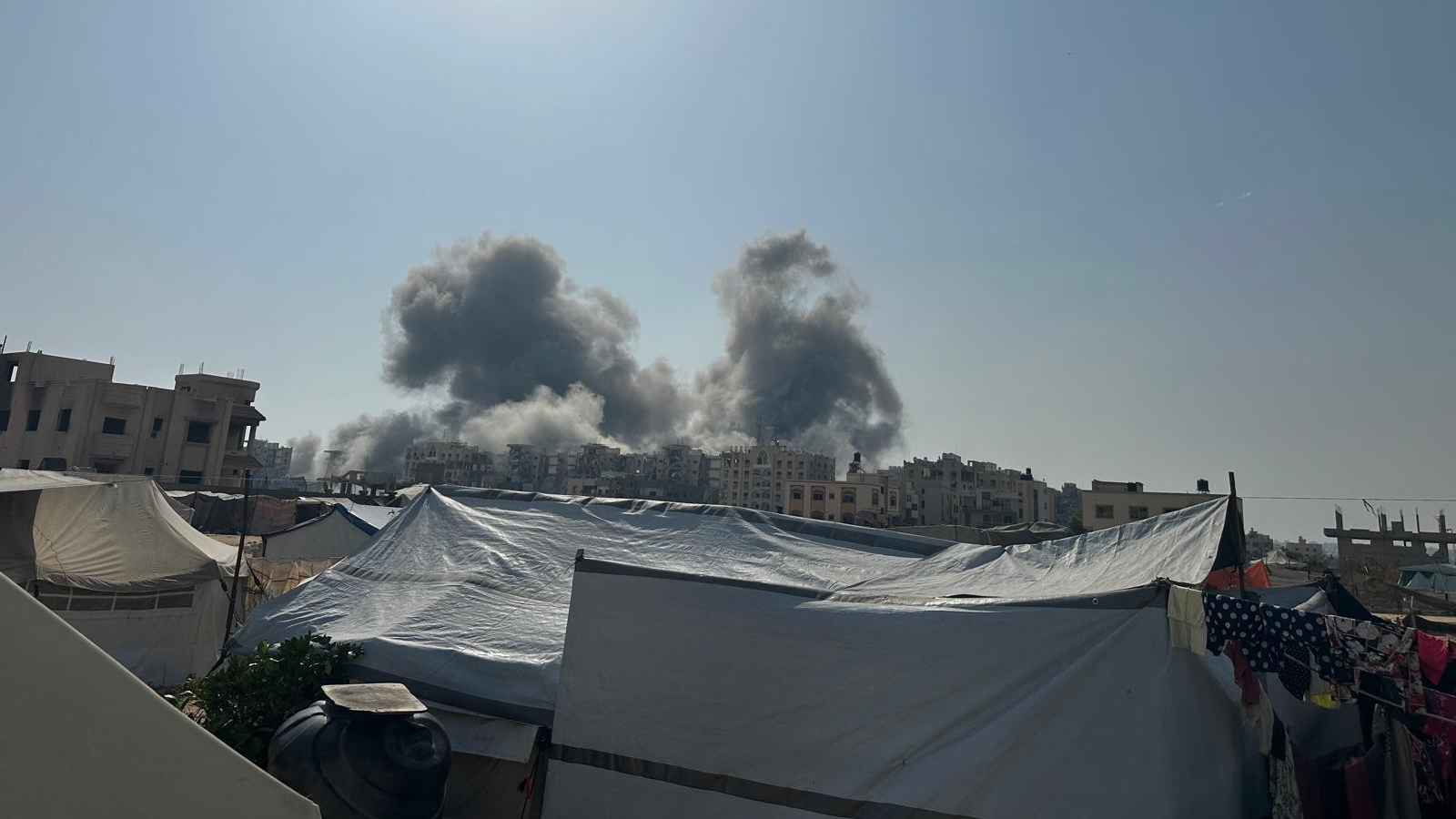 Smoke rises from buildings, tents can be seen in the foreground and a clothes line with washing hanging is in the corner.