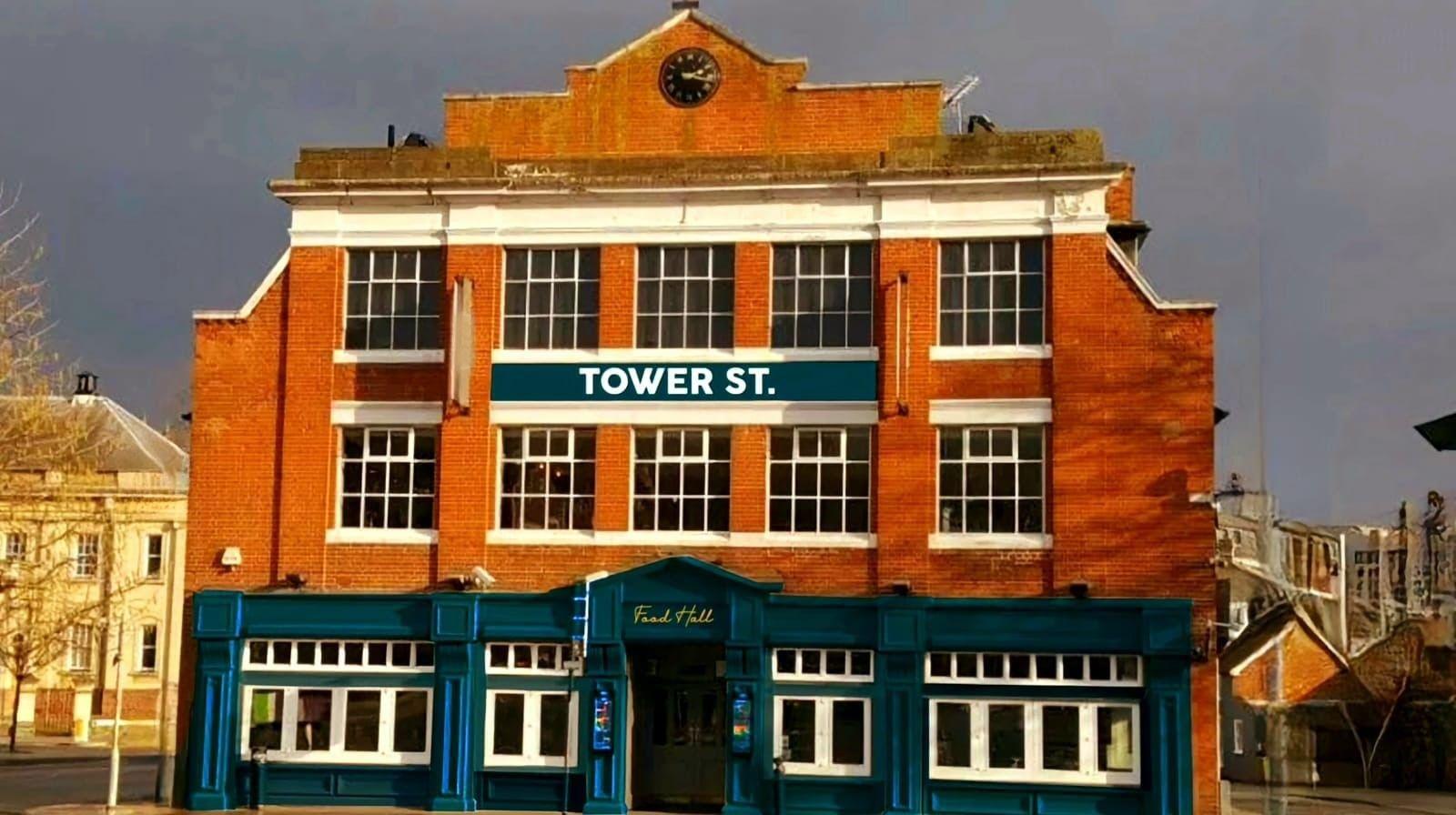 The exterior of the Tower Street Food Hall on Tower Street, Ipswich, a red brick three-storey building with blue frontage on the ground floor and white painted window frames on the first and second floors, a white sign saying "Tower St." between the first and second floors and a black clock above.