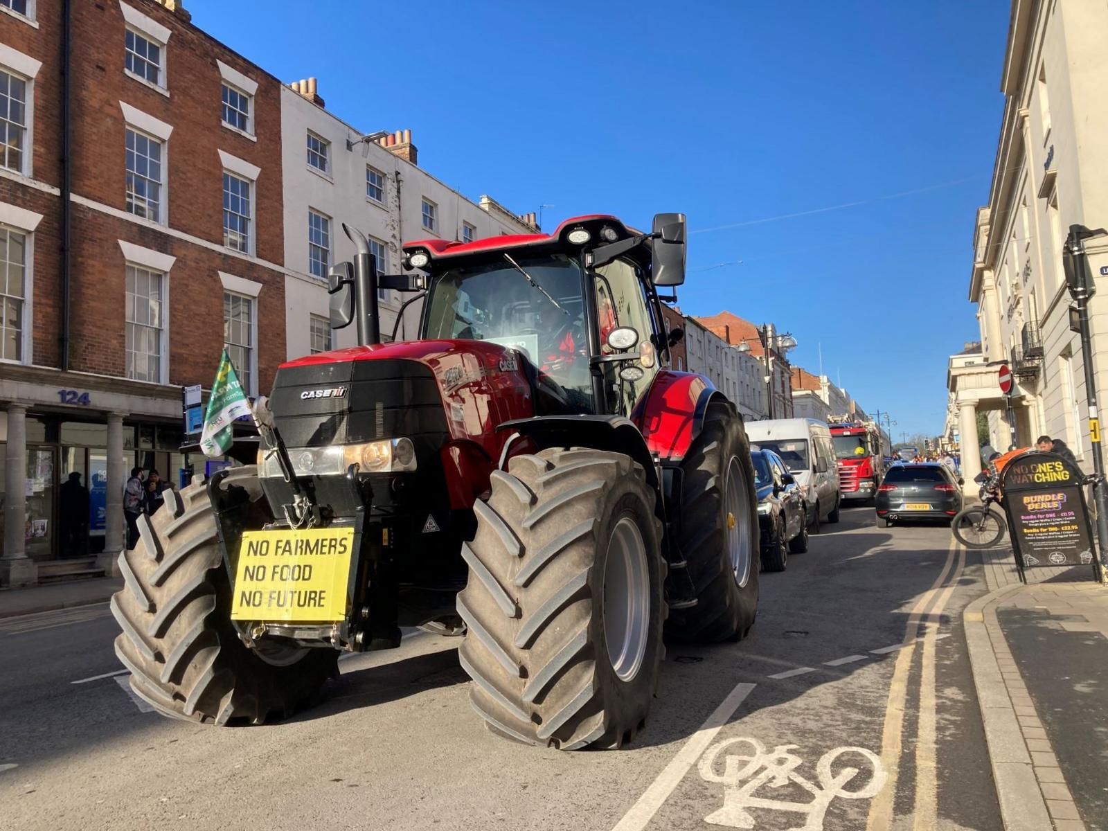 A red tractor driving down a high street on a sunny day. The tractor is decorated with a small green flag and a poster that says "NO FARMERS NO FOOD NO FUTURE".