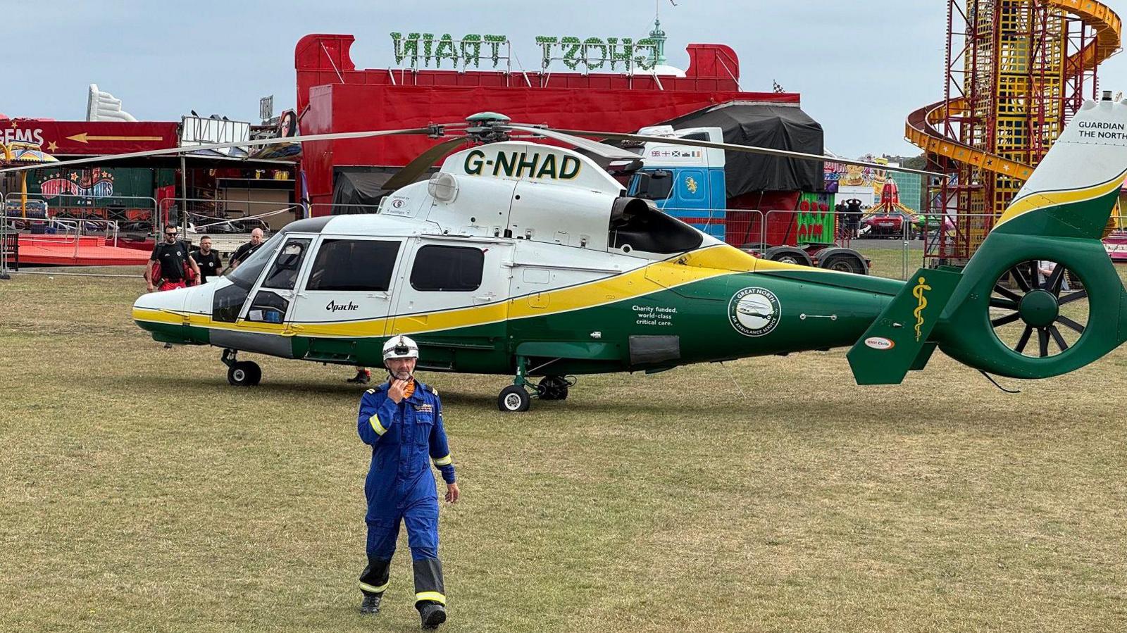 An air ambulance is landed on a grassed area in front of several fairground rides. A member of the emergency services can be seen in the foreground. He is wearing a blue uniform and white helmet.