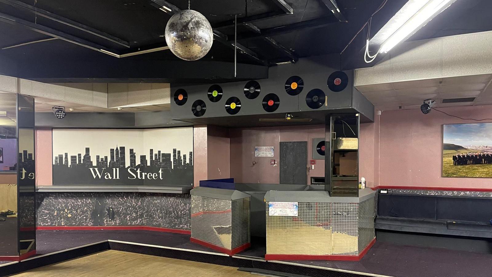 A mirror panelled DJ booth, with vinyl records stuck on the wall, stands next to an image of the New York skyline with the words 'Wall Street' on, the final name of the club before it closed