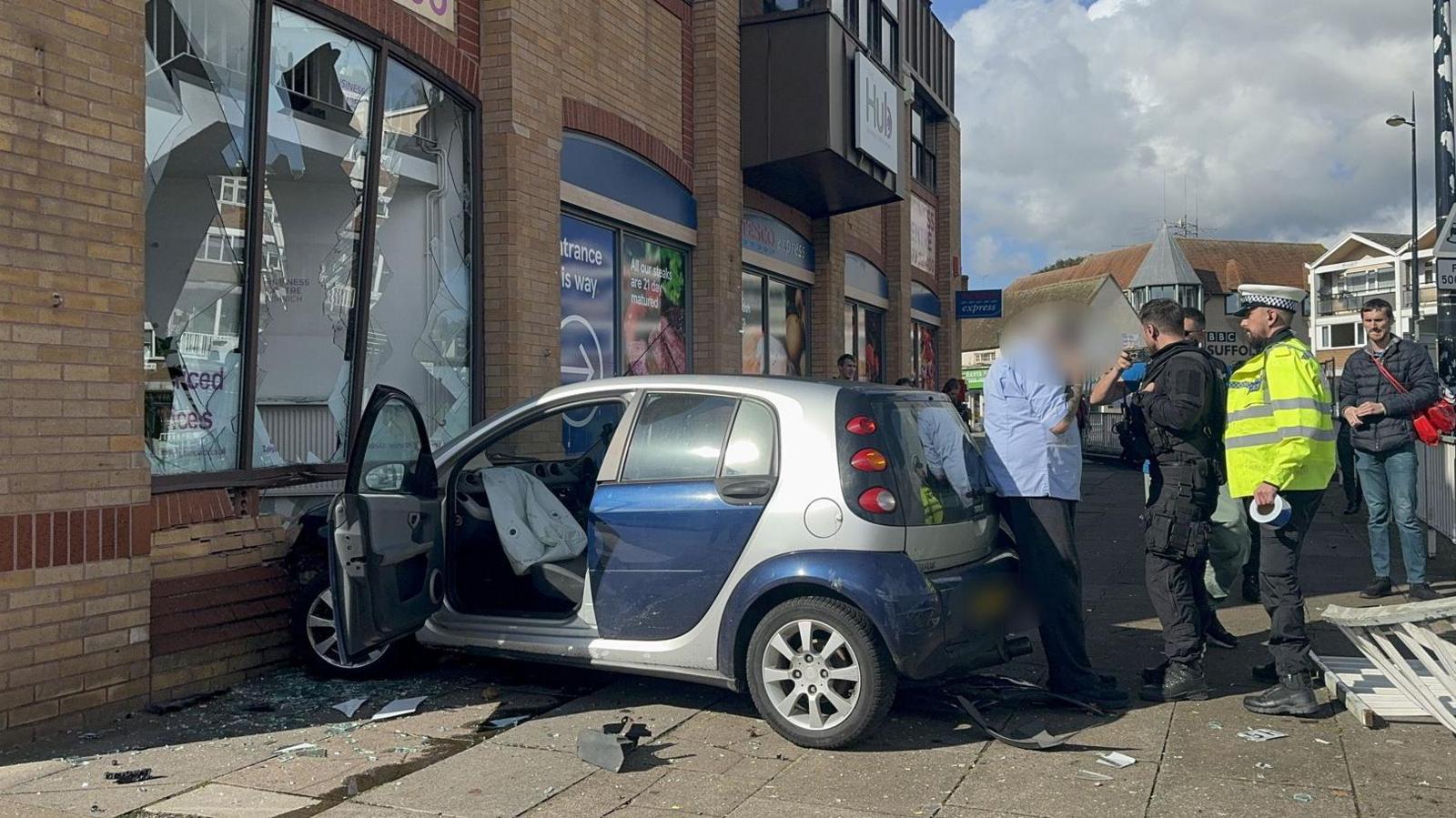 A navy Smart car crashed into a brick building with police officers and onlookers surrounding the scene