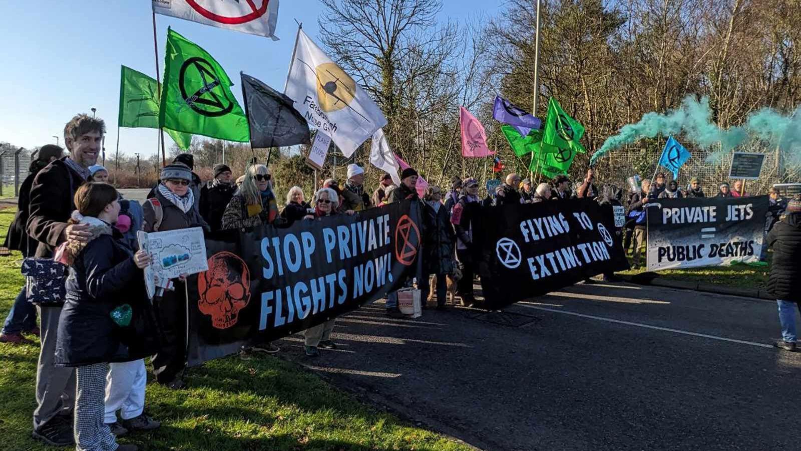 Campaigners with banners against the airport expansion standing outside the airport. The banners say stop private flights now, flying to extinction and private jets = public deaths.
