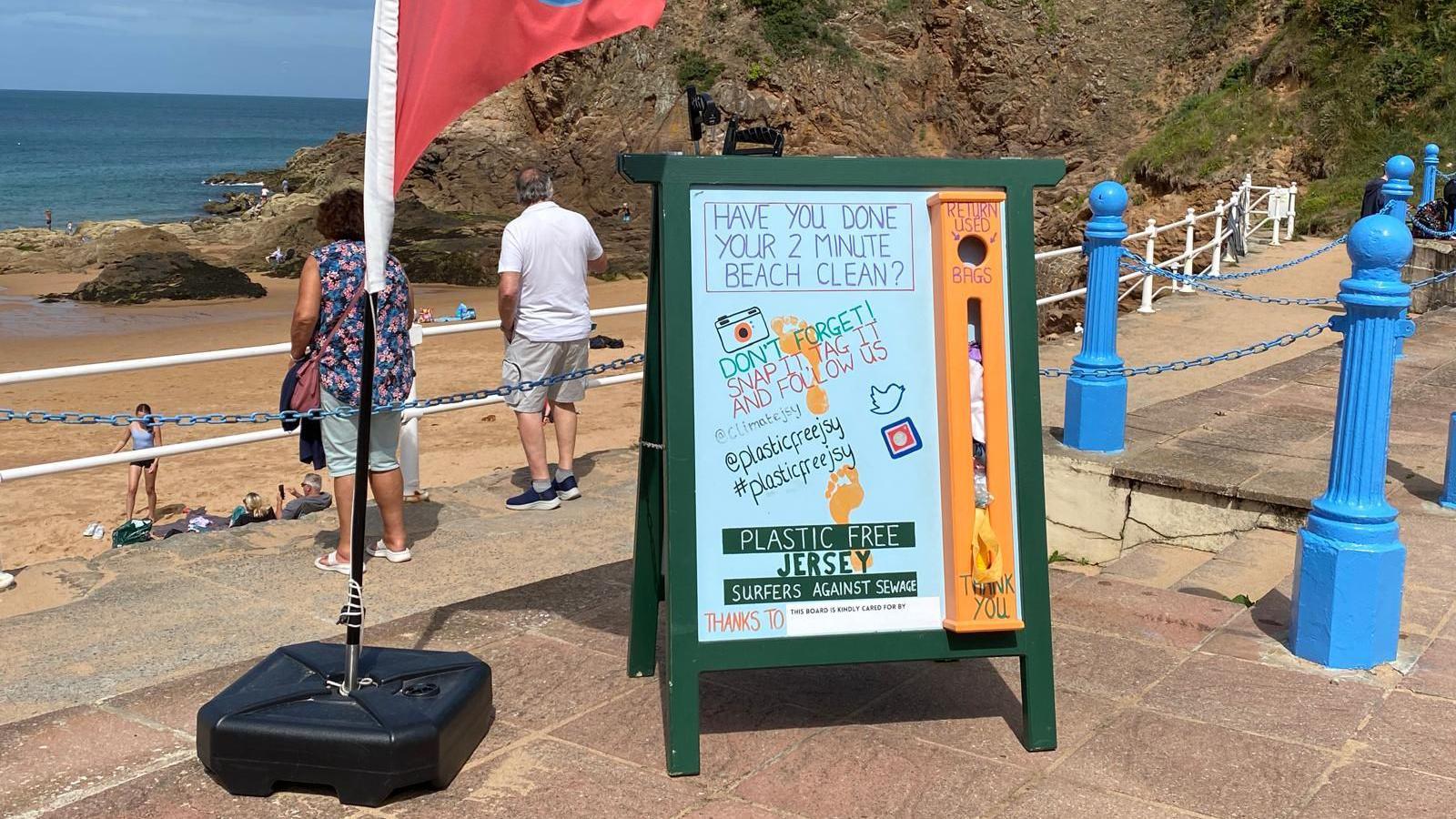 A sign saying 'Have you done your two-minute beach clean?' on a beach promenade. A couple are standing looking out to sea behind the sign and there are cliffs to the right of the image