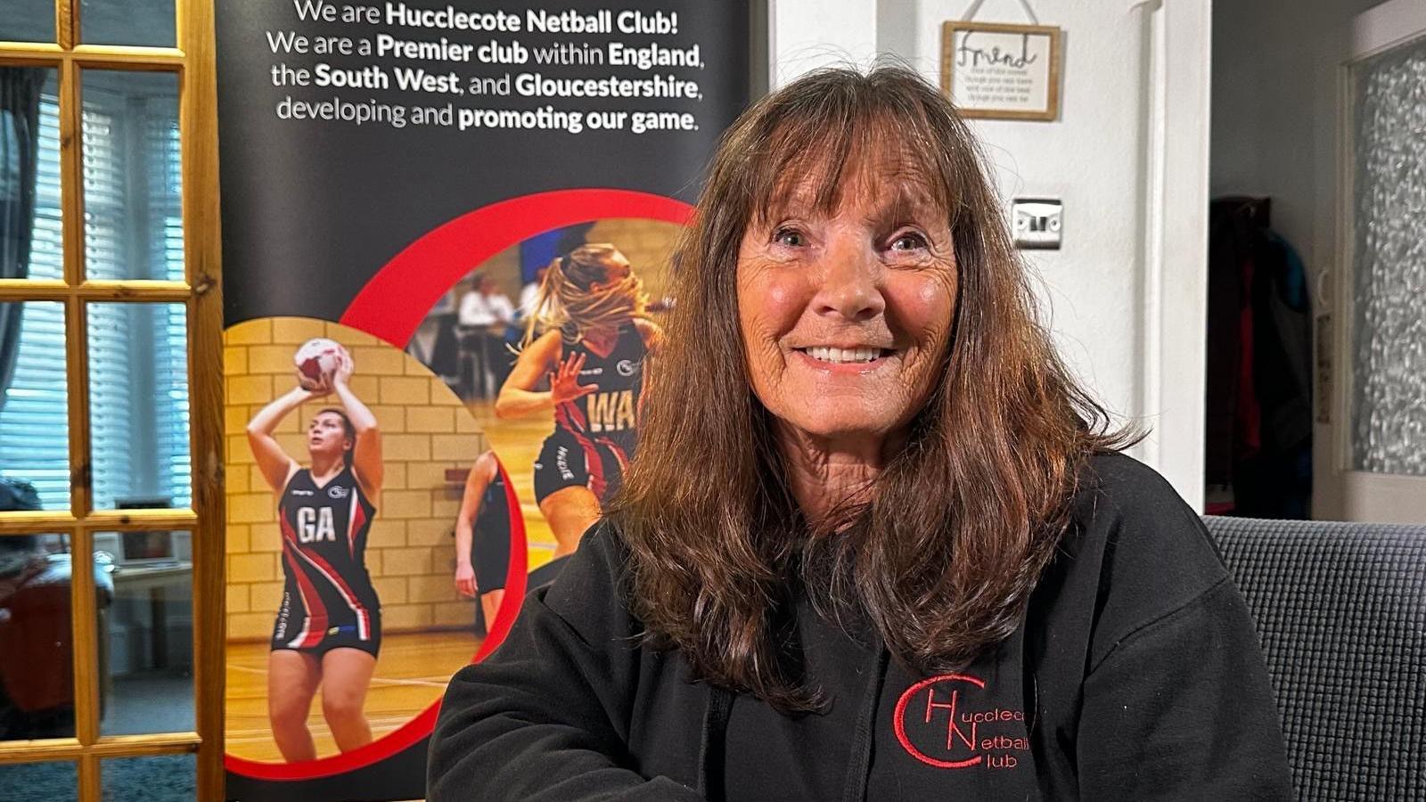 Sue Wainwright sitting at home, in front of a banner which reads "we are Hucclecote Netball Club, we are a premier club within England, the south west and Gloucestershire." She is wearing a black top