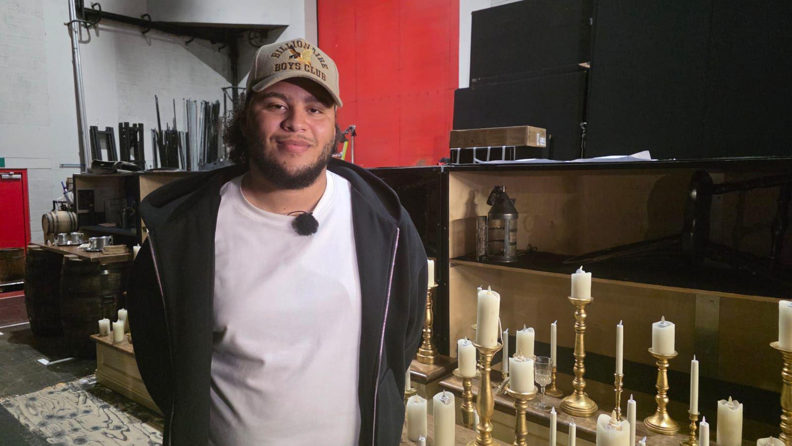 A picture of a man stood in front of a camera smiling. He has a black beard and is wearing a cream hat. He is wearing a white shirt and a black jacket. He is stood in front of a set with a number of candles. 