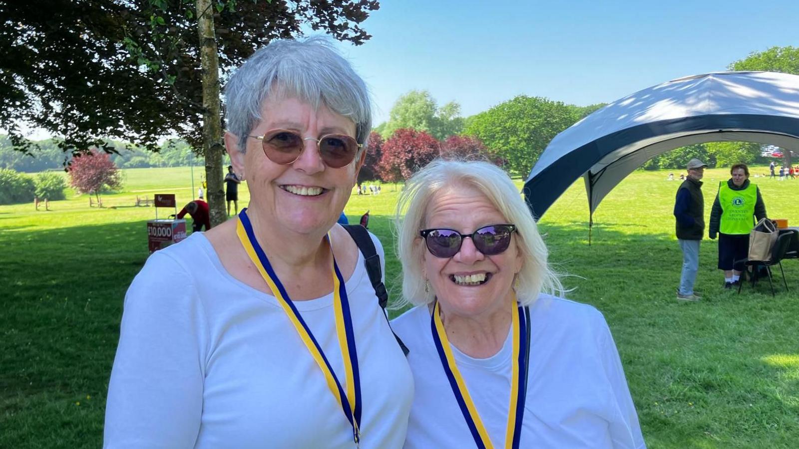 Two women with grey and white hair smile at the camera. They are both wearing white t-shirts and sunglasses. 