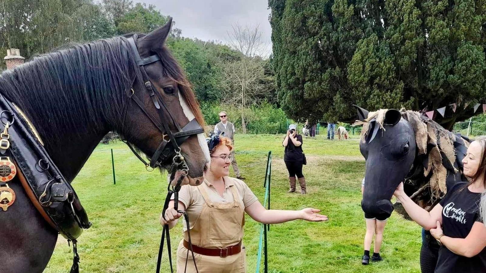 A woman in beige dungarees holds the reins of a large shire horse and a puppet horse held by a woman wearing a black top. Other people are watching and taking photos in a green field lined by trees.