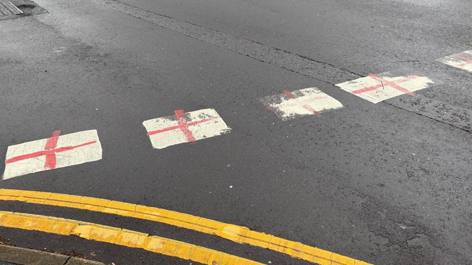 A close-up of a grey tarmac road, with bright double yellow lines in the foreground. In the centre of the frame are white give way road markings, which have been covered with red crosses.