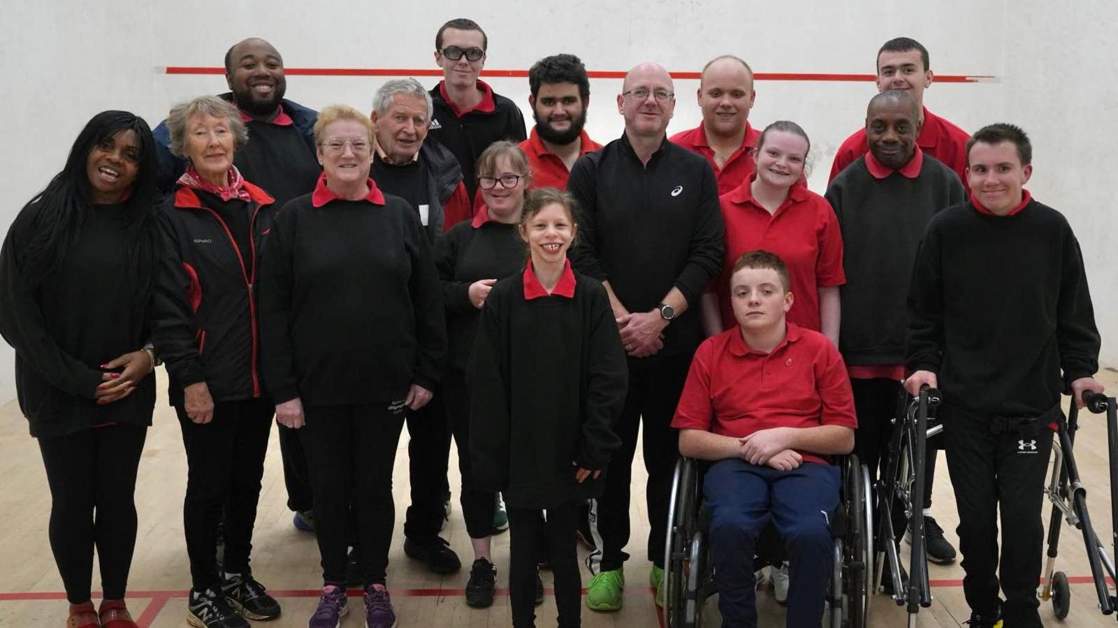 Some of the Orwell Panthers Athletics Club gathered together in a sports hall. They are standing in a group and smiling at the camera.They are wearing a mixture of their club sports kit including black jumpers and red t-shirts.