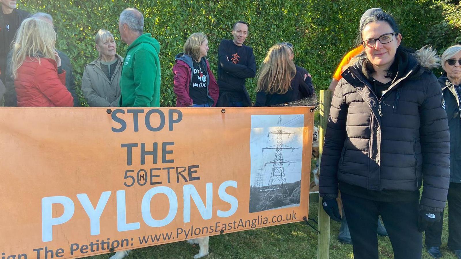 A photo of Jenny Keen. She is standing by the side of a protest sign. She is wearing a black puffer coat with black gloves and black leggings. She has black hair and is wearing black gasses. Other protesters can be seen behind her.