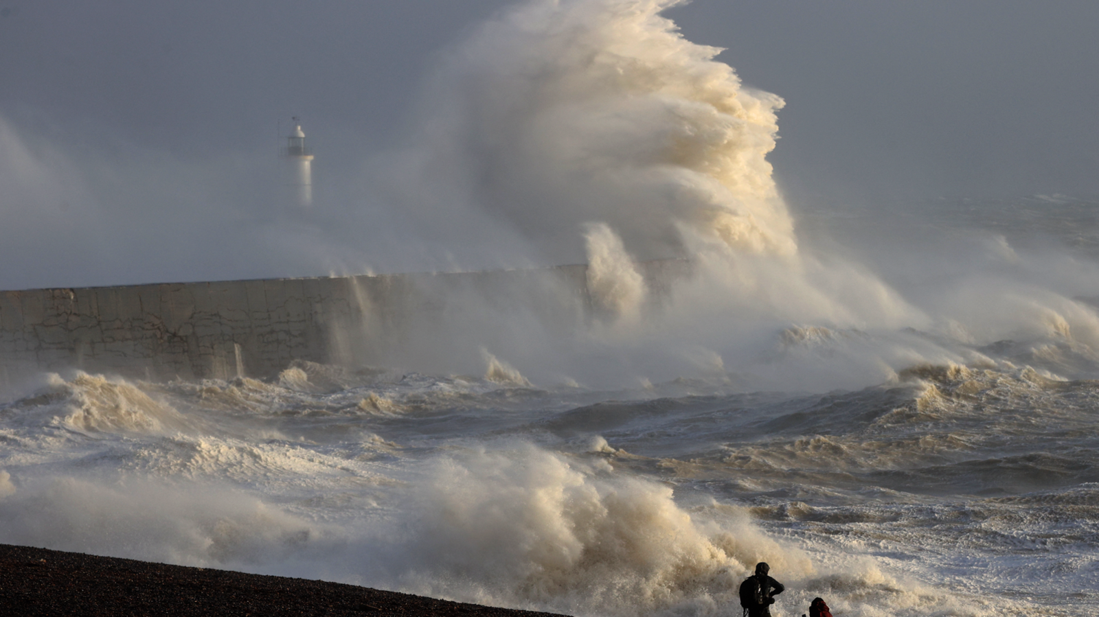 People watch as waves crash over a breakwater with a lighthouse in the distance