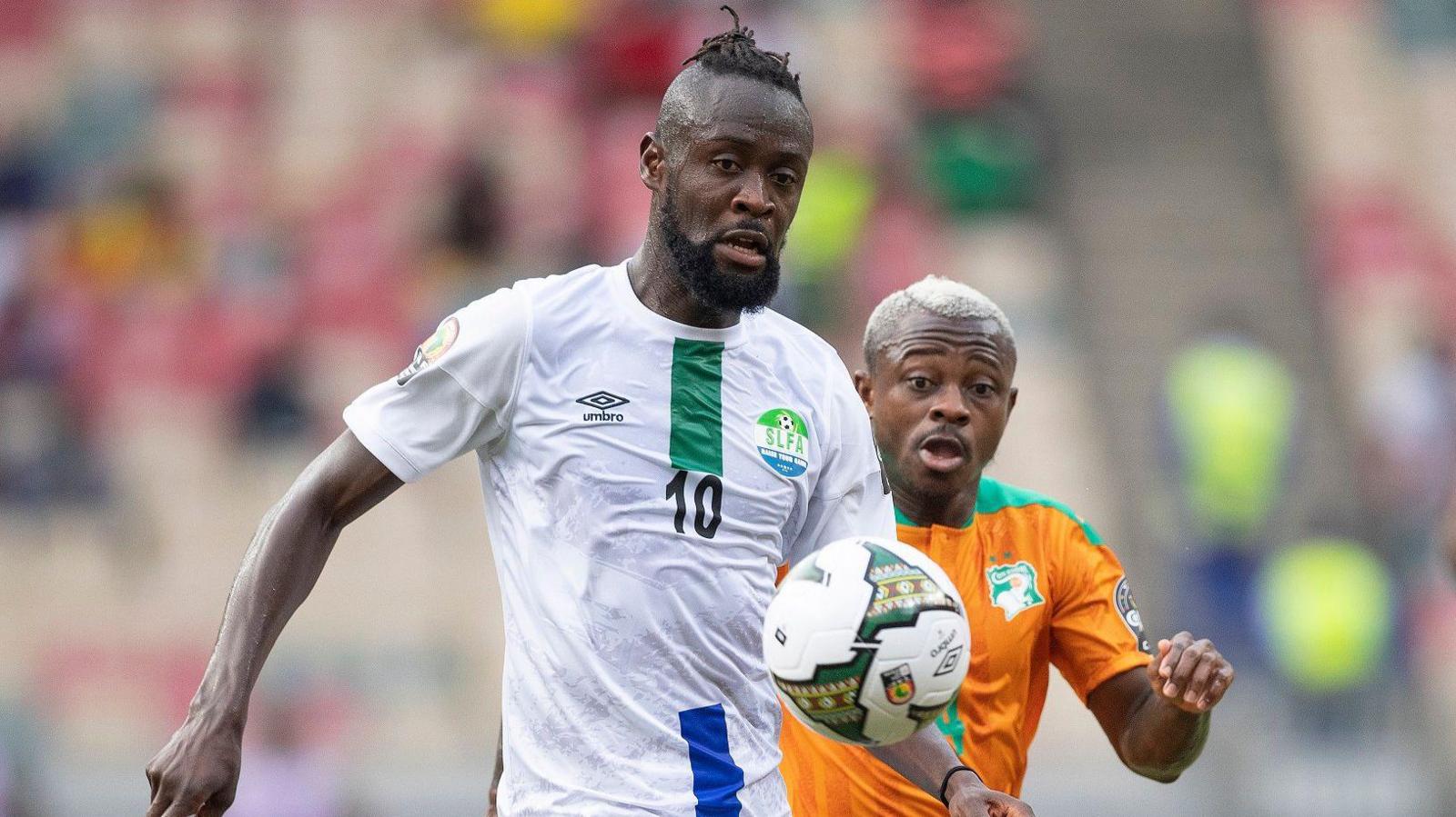 Kei Kamara of Sierra Leone in a white shirt challenges for a bouncing ball against Ivory Coast's Jean Michael Seri in an orange shirt at the 2021 Afcon. 