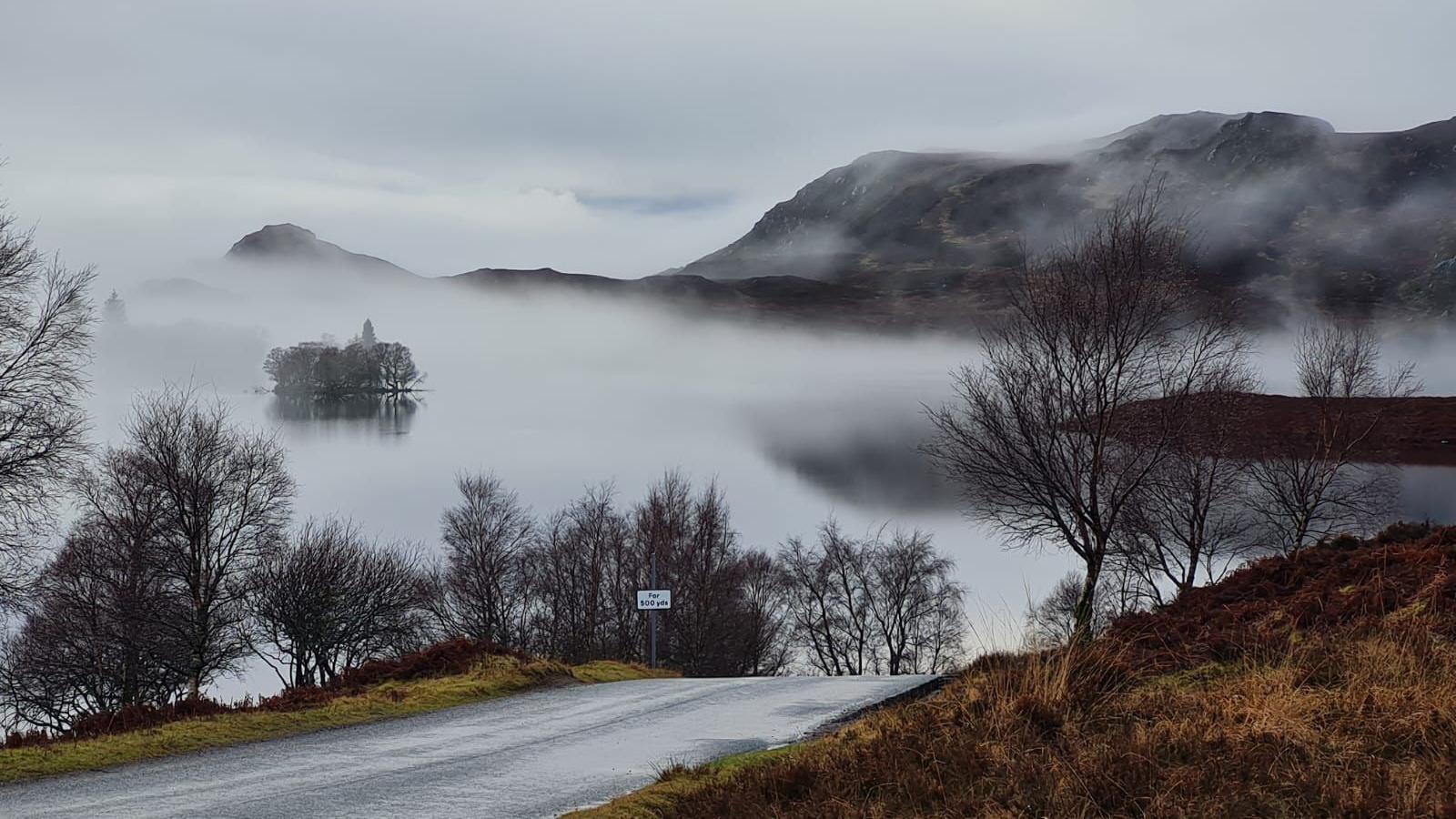 A lake is hidden by fog. A road emerges out of the fog with bare trees on each side. Hills are seen in the background and it's a cloudy day. 