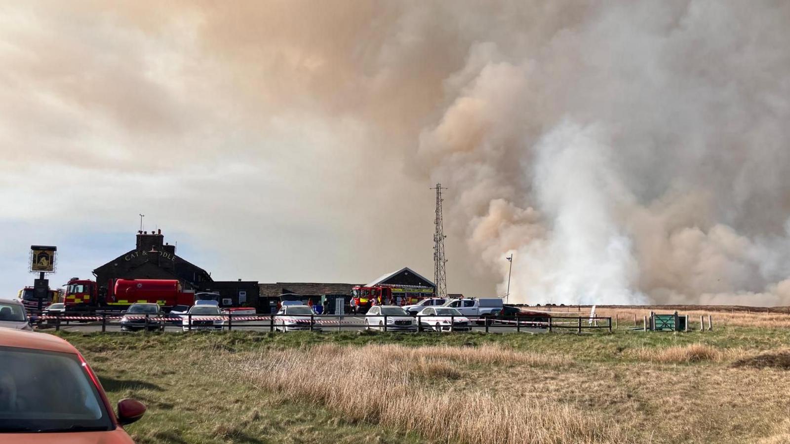Smoke fills the sky behind the Cat and Fiddle distillery. Fire engines are parked in the car park and a cordon is in place.