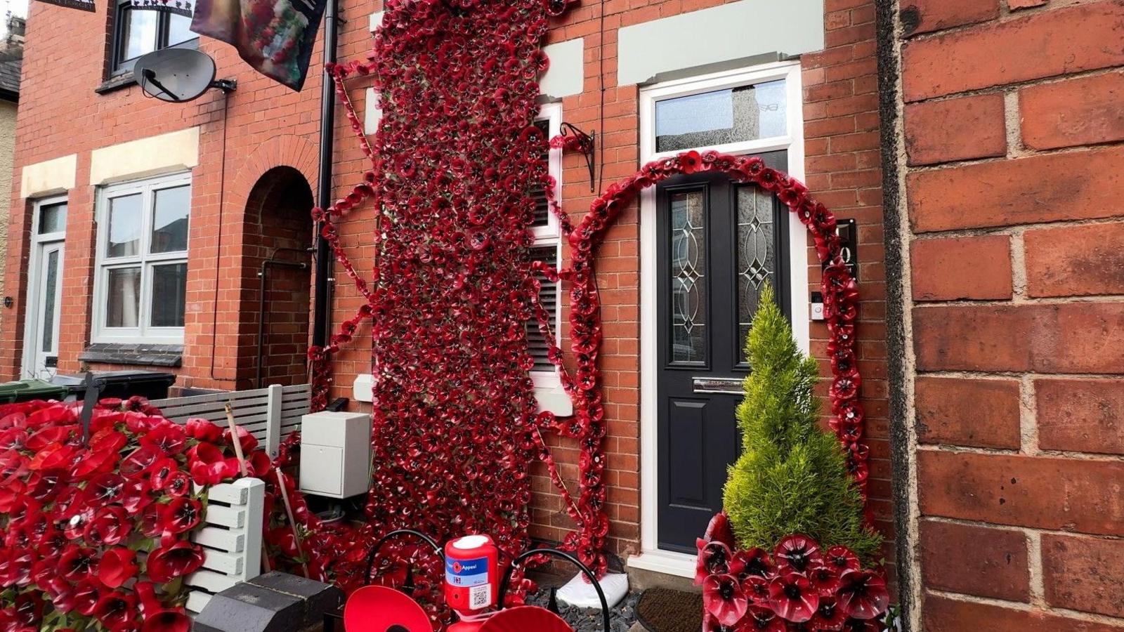 A terraced house, with red poppies hanging from the top window. An arch is on display around the front door, and a Royal British Legion collection box can be seen on the front gate.