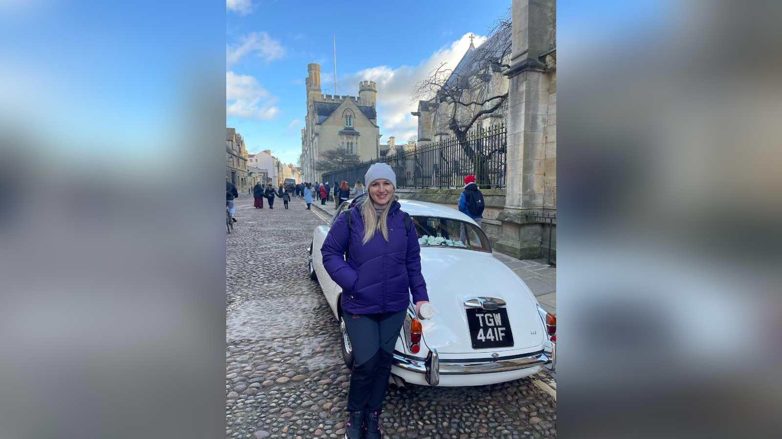 A woman stands in front of an old-fashioned white car outside a Cambridge University college 