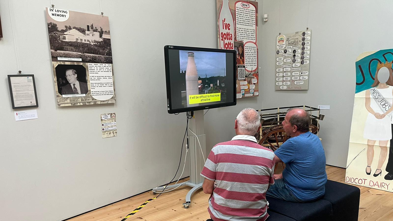 Two men are watching archive footage on a TV in an exhibition room. They are sat down beside other memorabilia. Placards surround the TV on the wall.