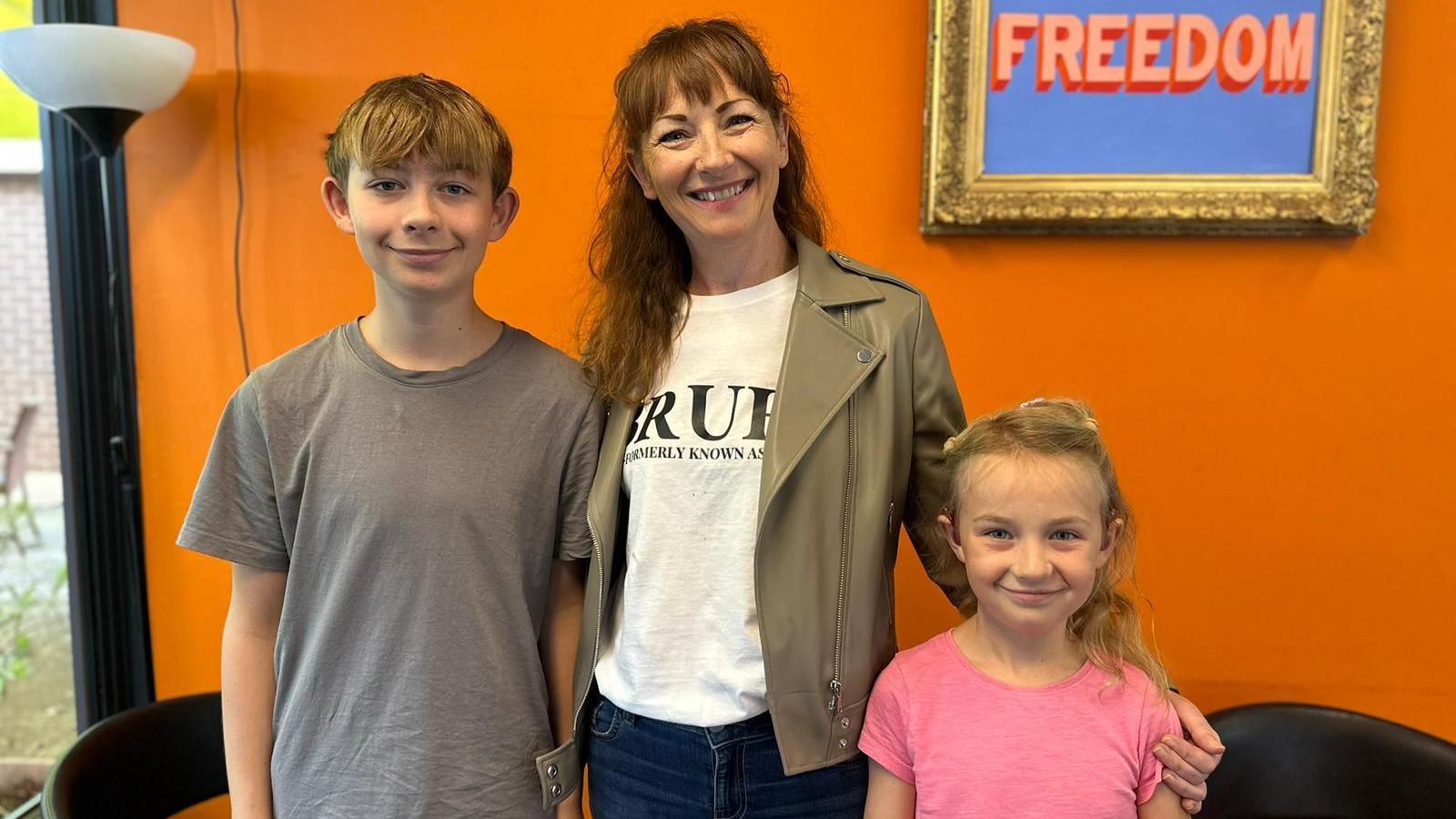Coletta Todd with long ginger hair and a fringe standing between her 14-year-old son henry who is wearing a grey t shirt and her sweven-year-old daughter Mary who is wearing a pink t-shirt. They are standing in front of a bright roange wall with a lamp to one side and gold frame on the wall with the words Freedom in typographical art