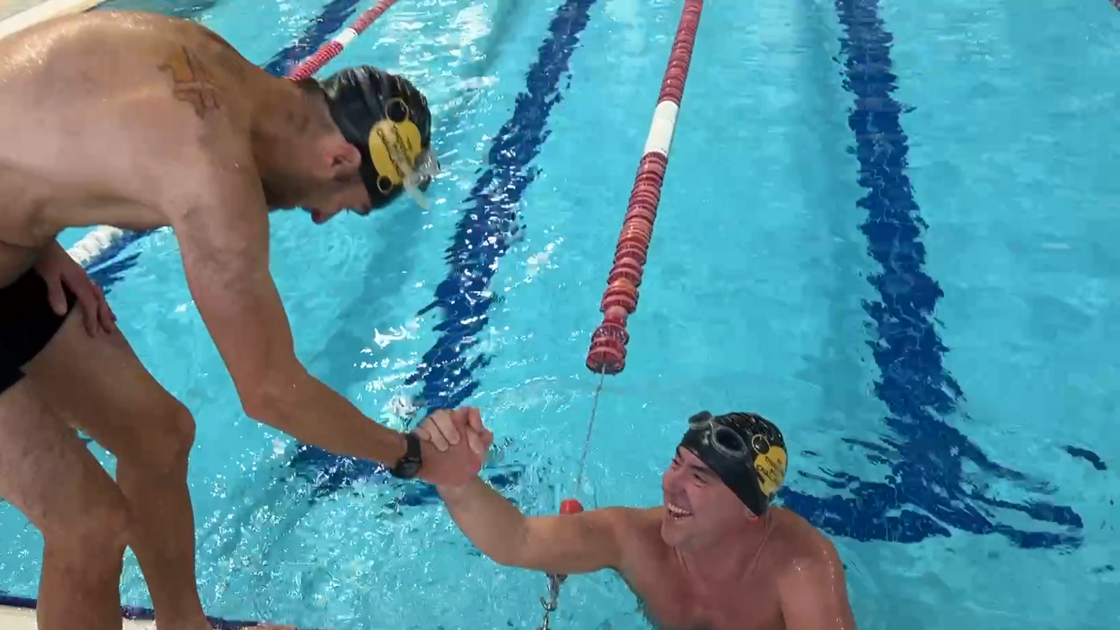 Edward Sault, a man in a swimming pool wearing goggles and a swimming cap, reaches up to shake hands with Steve Harris, a man wearing black swimming shorts, who is standing at the side of the pool.