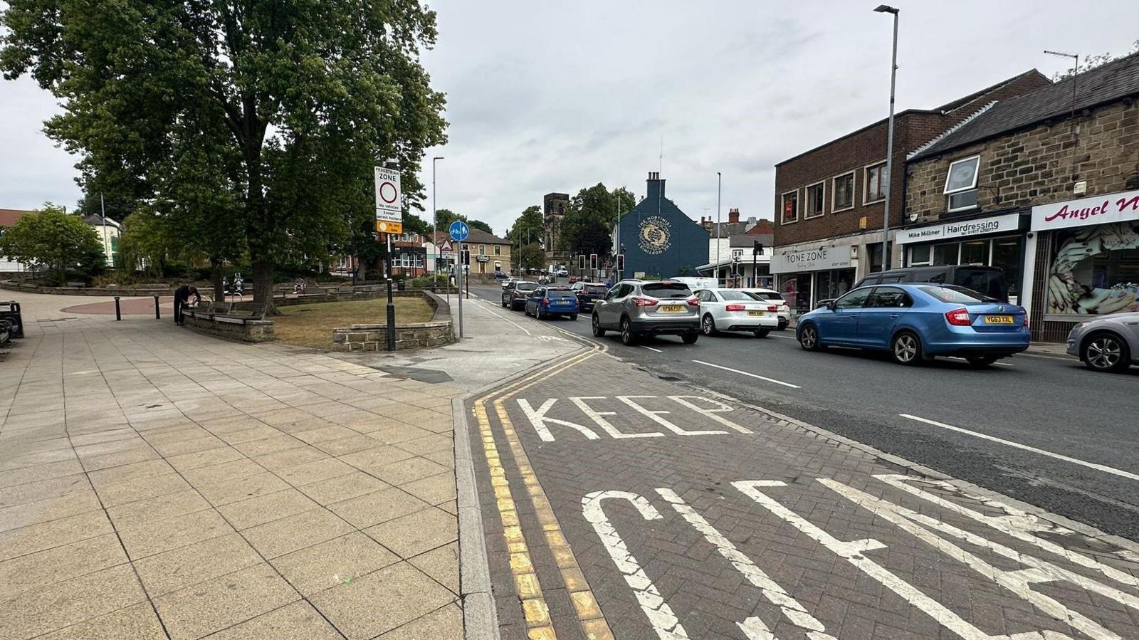 A road with two lanes of cars travelling on it. An area of the road is marked 'Keep Clear'. A paved pedestrian area on the left side with a small patch of grass and a tree. A row of shops on the opposite side of the road.